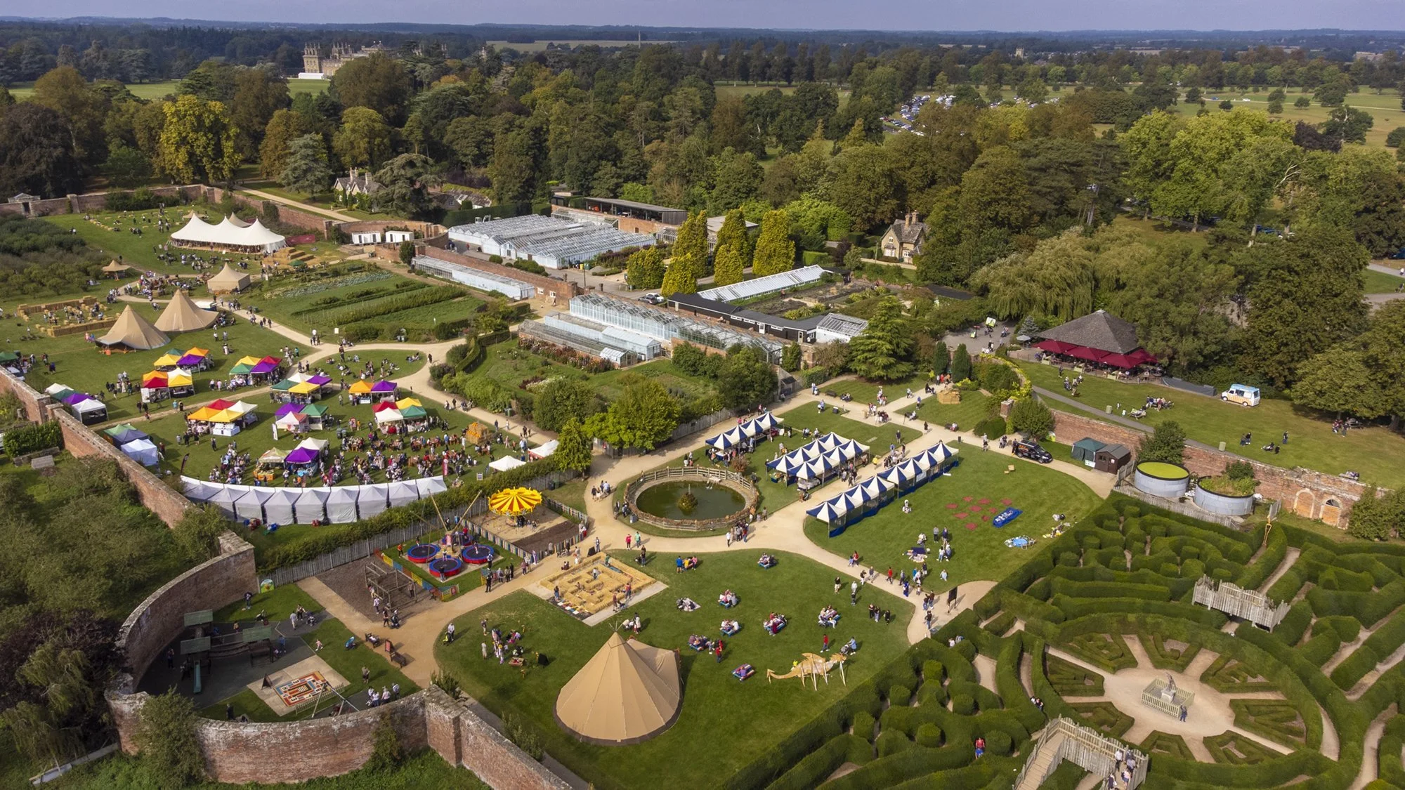 Aerial view of a large outdoor event with tents, greenhouses, a carousel, picnic areas, and a garden, surrounded by trees and open fields.