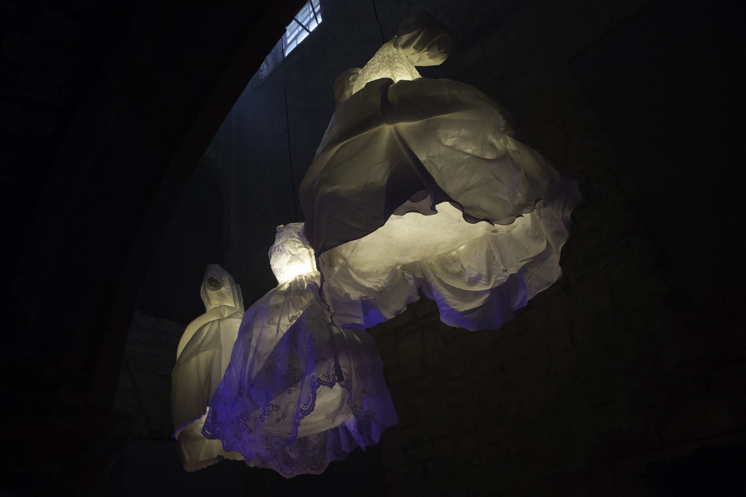 Illuminated abstract textile sculpture hanging from the ceiling in a dark room, featuring flowing fabric with lace and embroidered details.