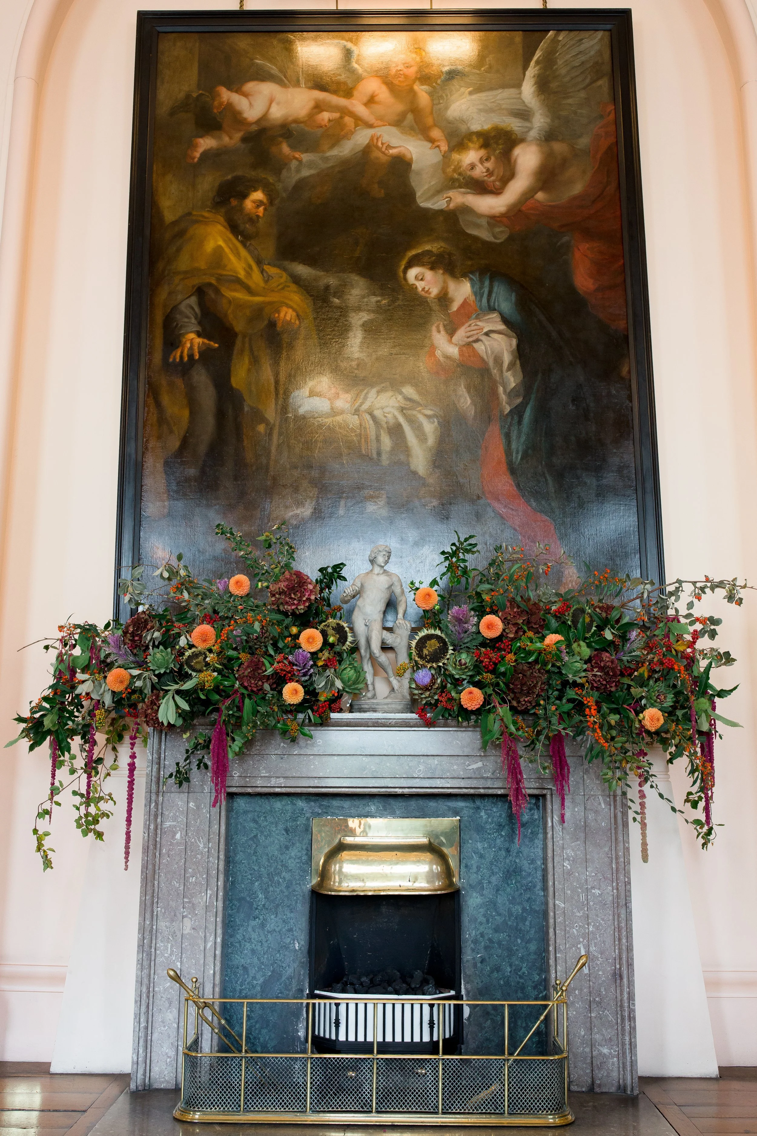A large fireplace decorated with a floral arrangement, a small statue, and a large painting depicting a biblical scene with angels and a woman looking down at a baby in a manger.