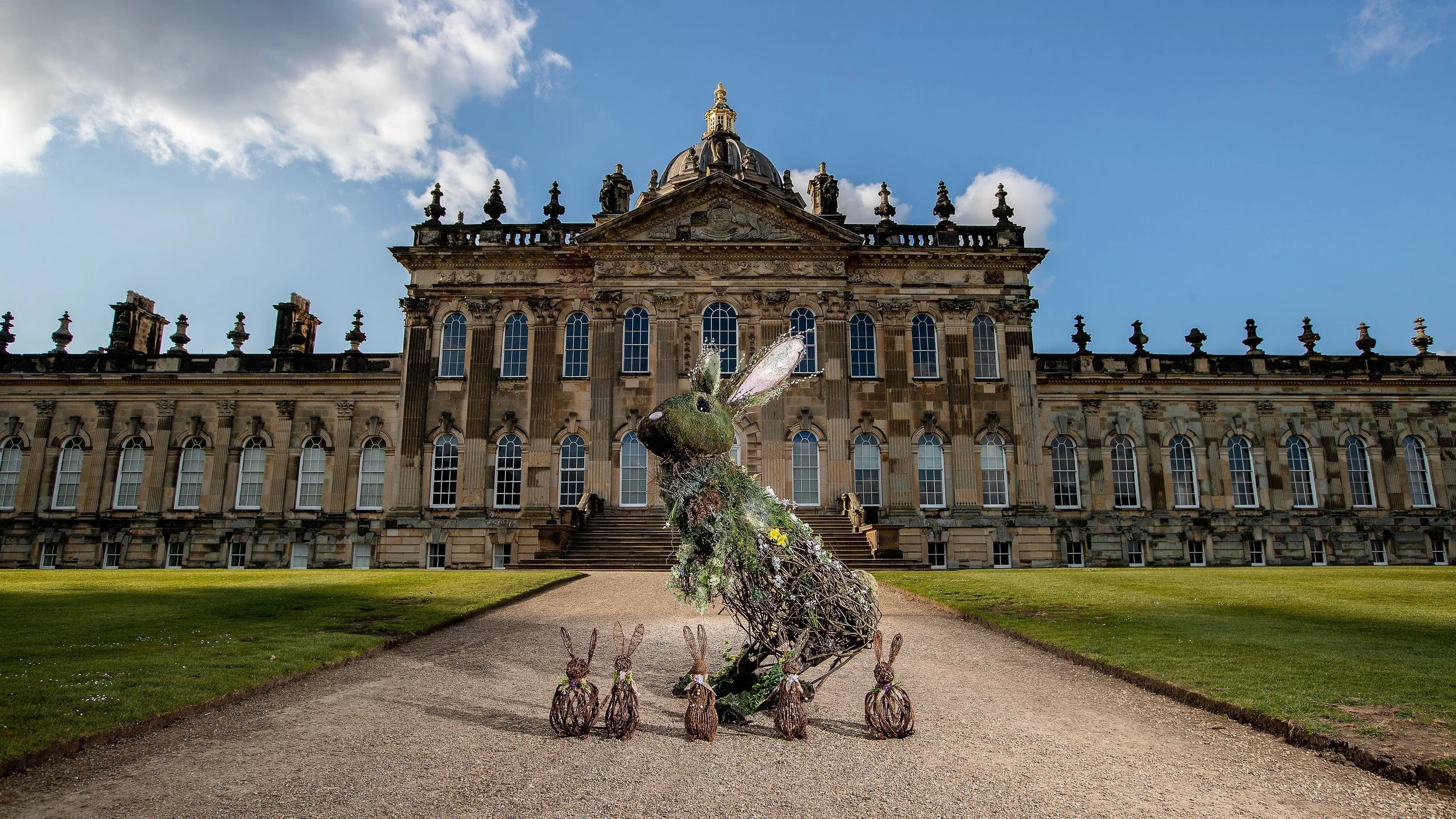 A large historic building with ornate architecture, multiple windows, and a domed roof. In front, there are decorative bunny statues and an elaborate sculpture of a rabbit made of plants and branches.