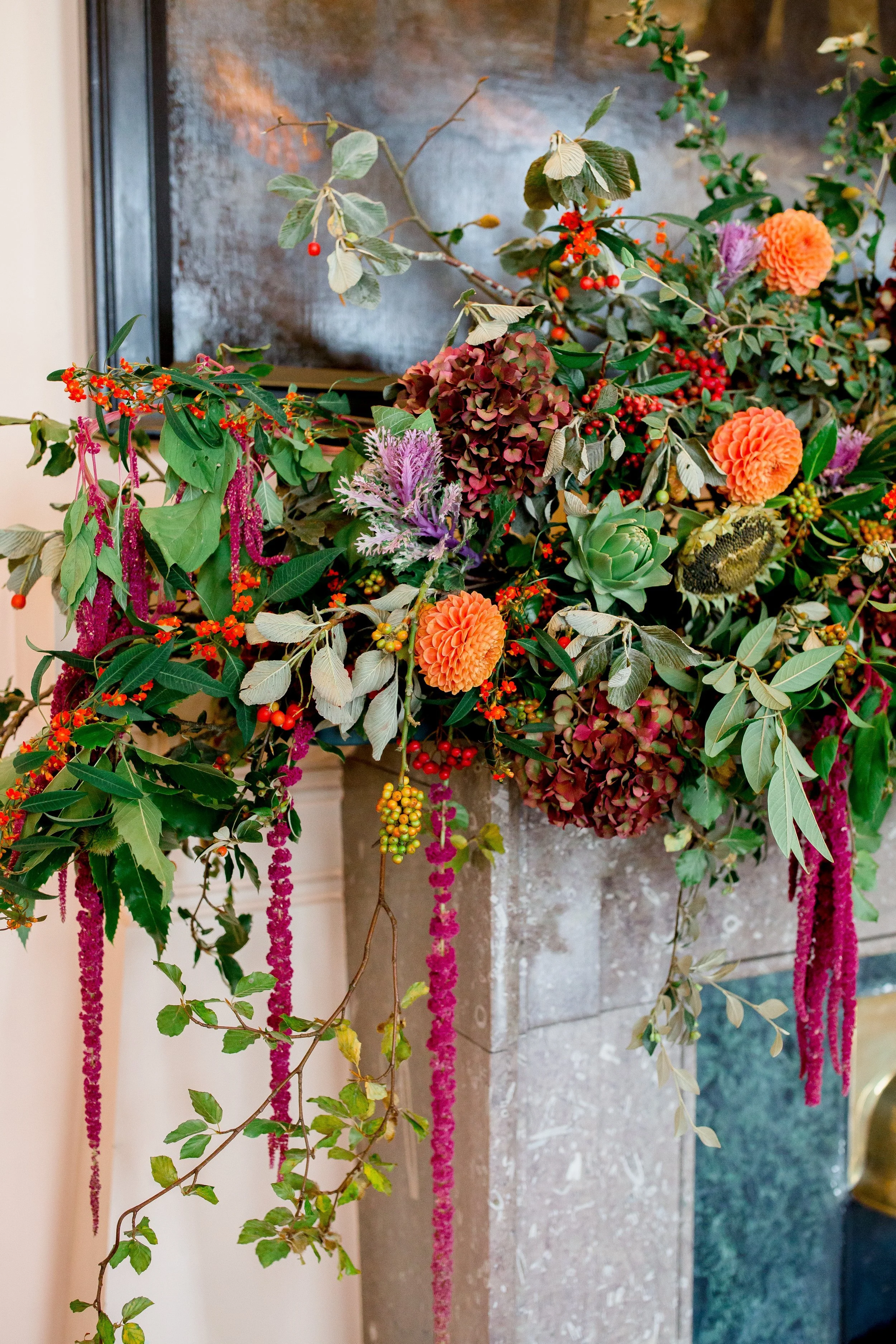 A floral arrangement with orange dahlias, purple thistles, red berries, and various green leaves, placed on a stone pedestal with a dark background.