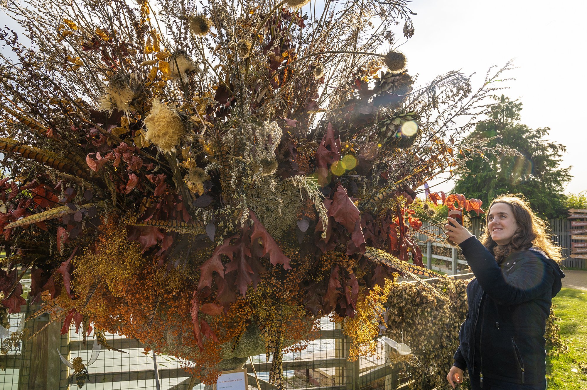A woman with curly hair and a black jacket is holding a phone and taking a picture of a large decoration made of dried flowers and leaves outdoors in sunlight.
