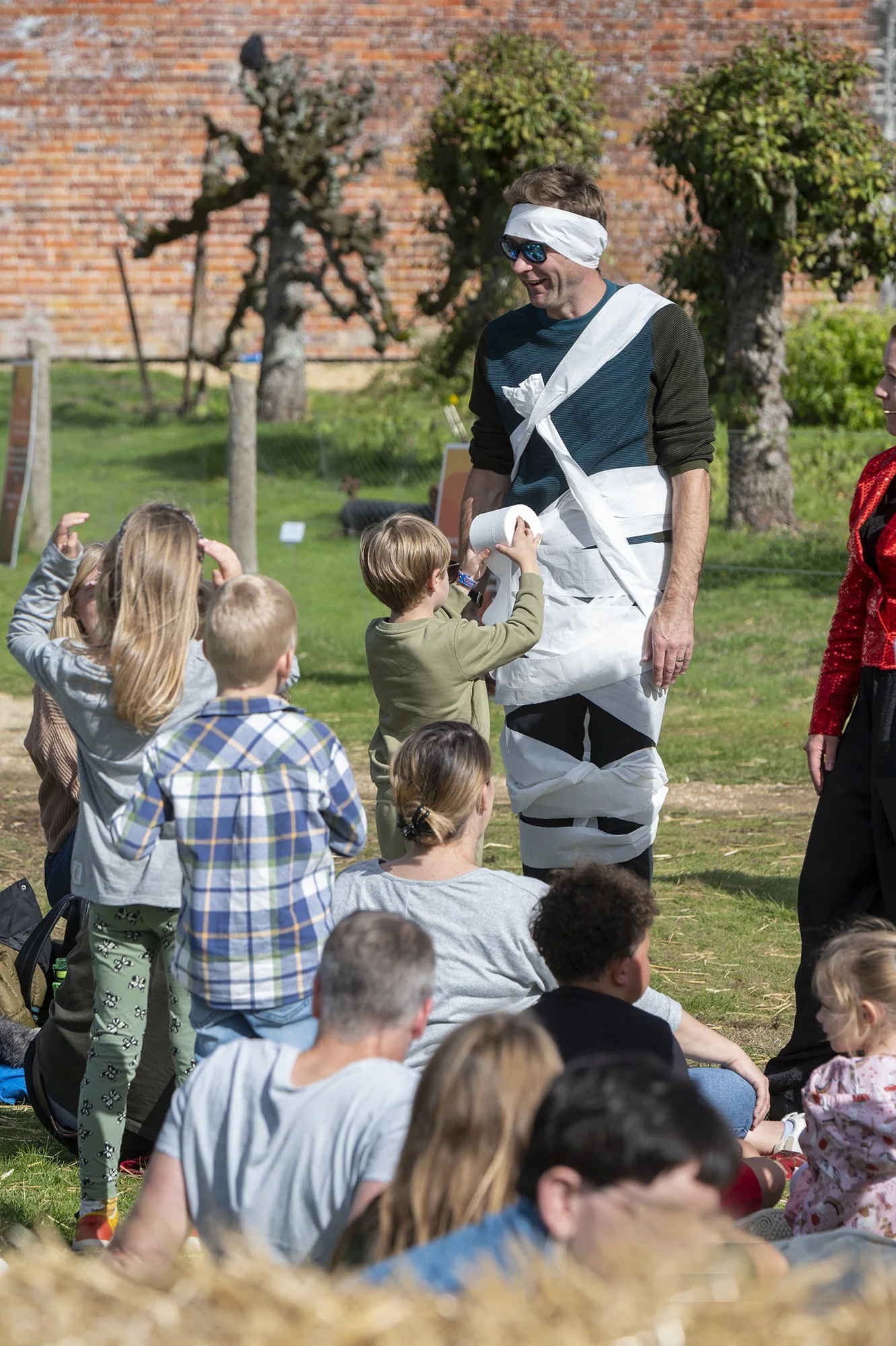 A man dressed as a mime or clown, wearing a diaper, sunglasses, and a white headband, interacting with a group of children outdoors. The children are sitting or standing on a grassy area with trees and a brick wall in the background.