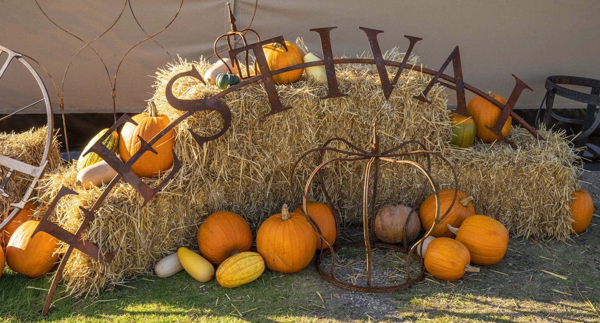 An autumn display with hay bales, pumpkins of various sizes, gourds, and decorative metal accents, set outdoors near water during daylight.