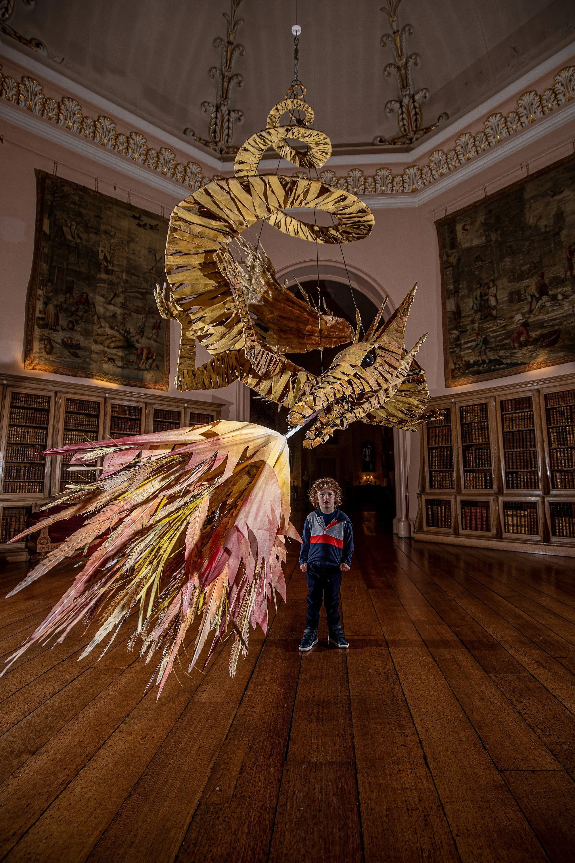A boy is standing in a gallery room with wooden floors and ornate decorations, facing a large, intricate sculpture resembling a mythical creature with wings, tail, and an elaborate headpiece, suspended from the ceiling.