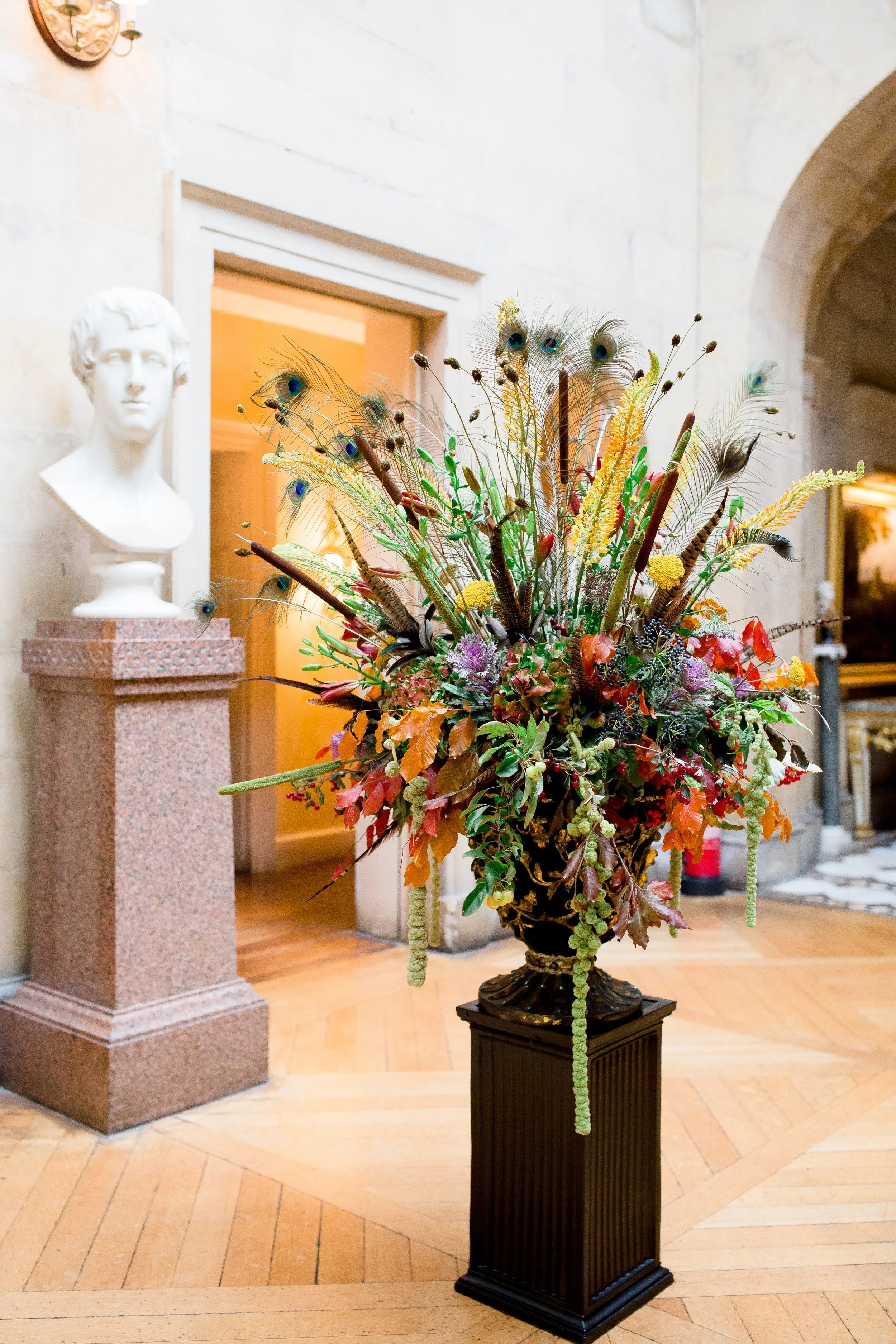A tall floral arrangement featuring peacock feathers, yellow, purple, and orange flowers, and green foliage in a black pedestal vase, situated inside a room with wooden flooring, stone walls, and classical bust statues.