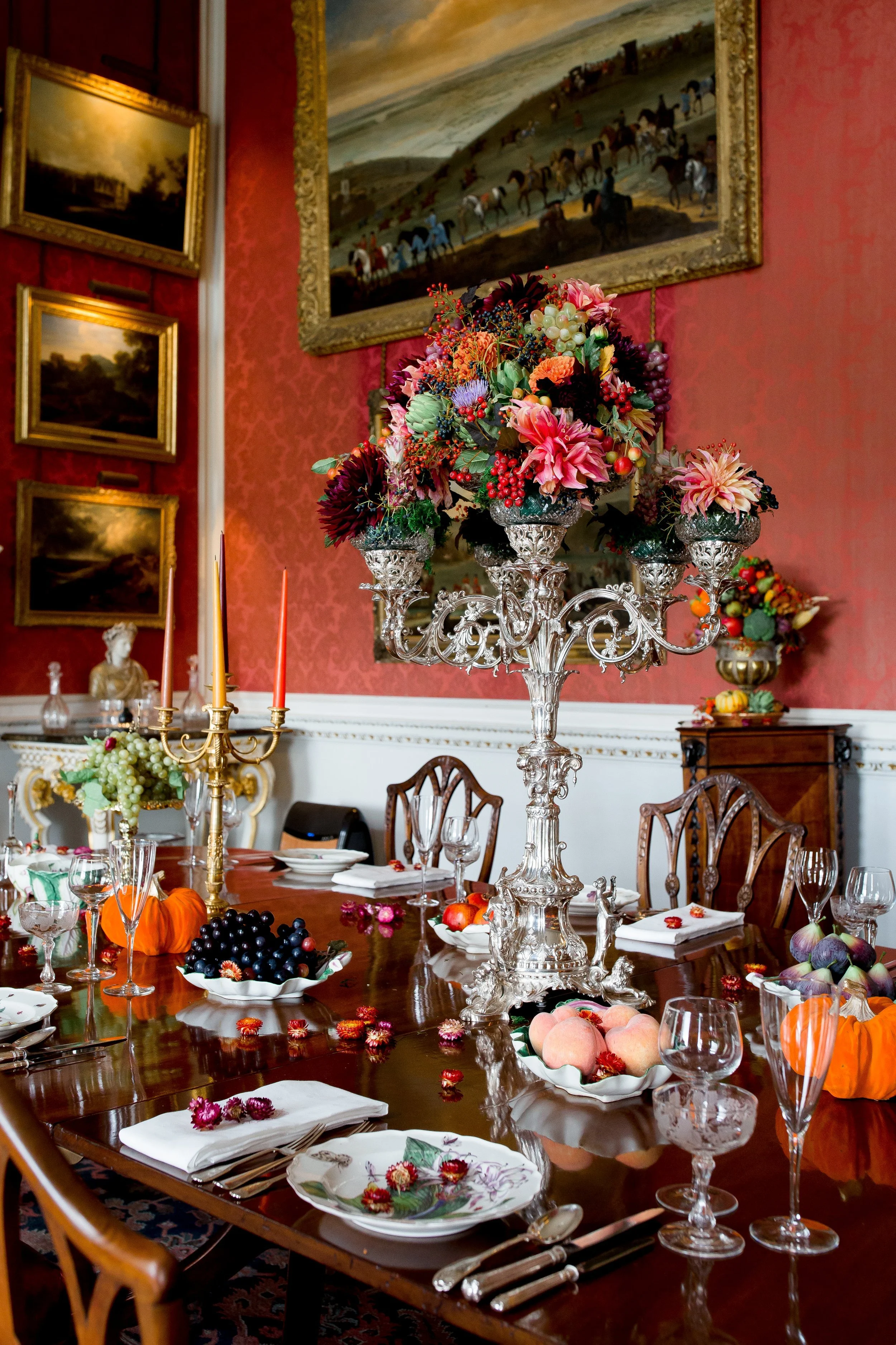 A decorated dining room set for a celebration with a large floral centerpiece, autumn-themed decorations, and arranged plates, glasses, and silverware on a wooden table. Framed paintings hang on red patterned walls.