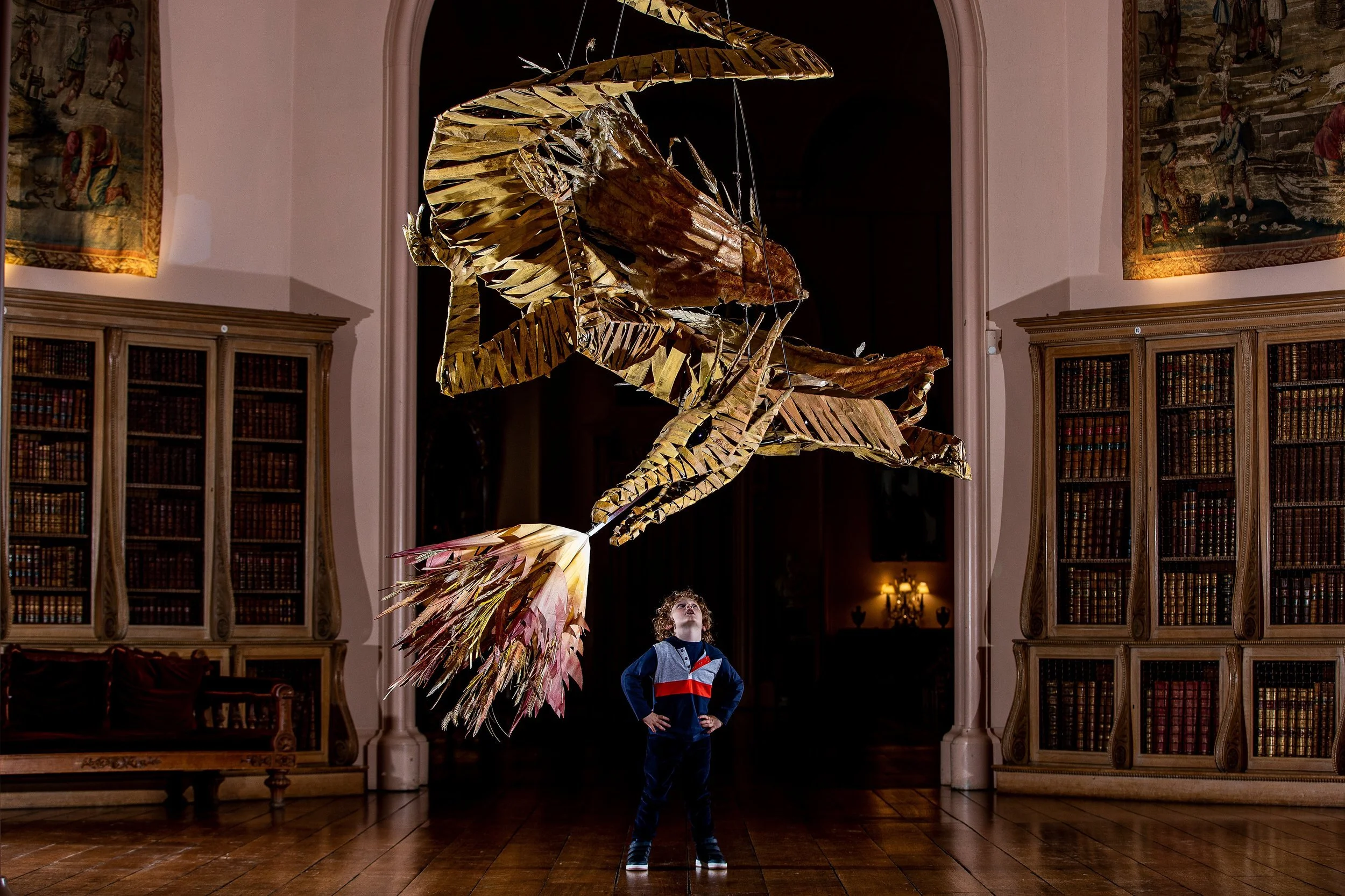 A young boy stands with hands on hips in a room with wooden floors and bookshelves, looking up at a large, intricate sculpture of a dragon made of metal and wood hanging from the ceiling.