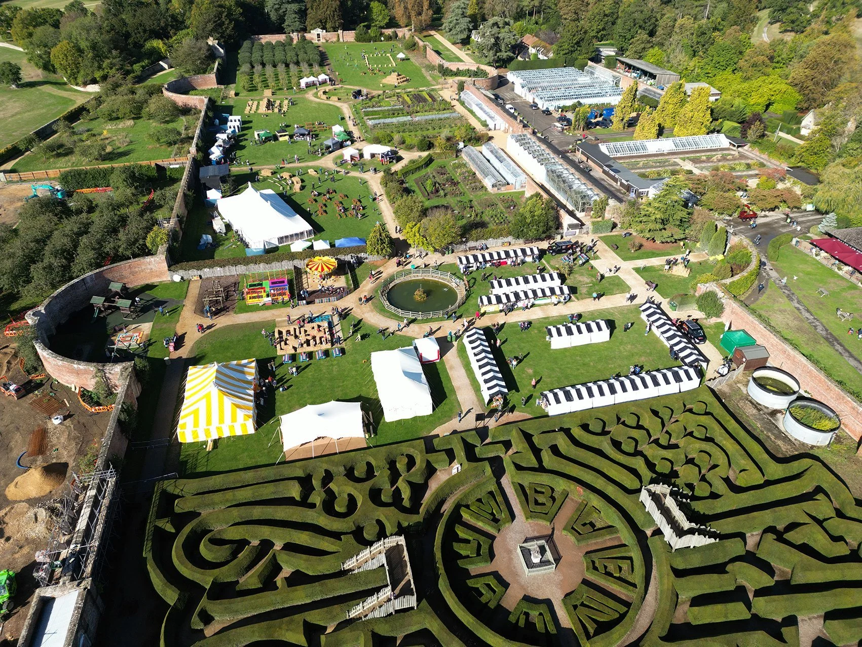 Aerial view of a garden festival with pathways, tents, greenhouses, a maze, and people walking, surrounded by trees and brick walls.