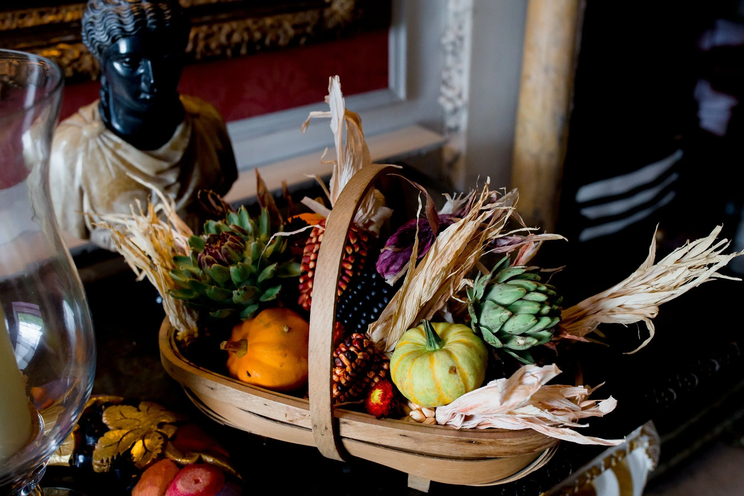 A basket filled with various fall-themed decorations and gourds on a table. A black mask with a gold cloth, a small white pumpkin, and colorful artificial flowers are visible among the decorations.