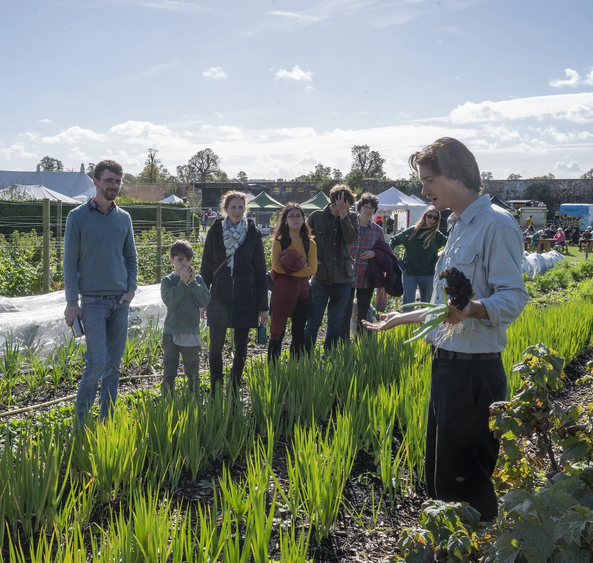 A man is speaking to a group outdoors at a community garden, with several people listening, surrounded by green plants and tents, under a blue sky with scattered clouds.