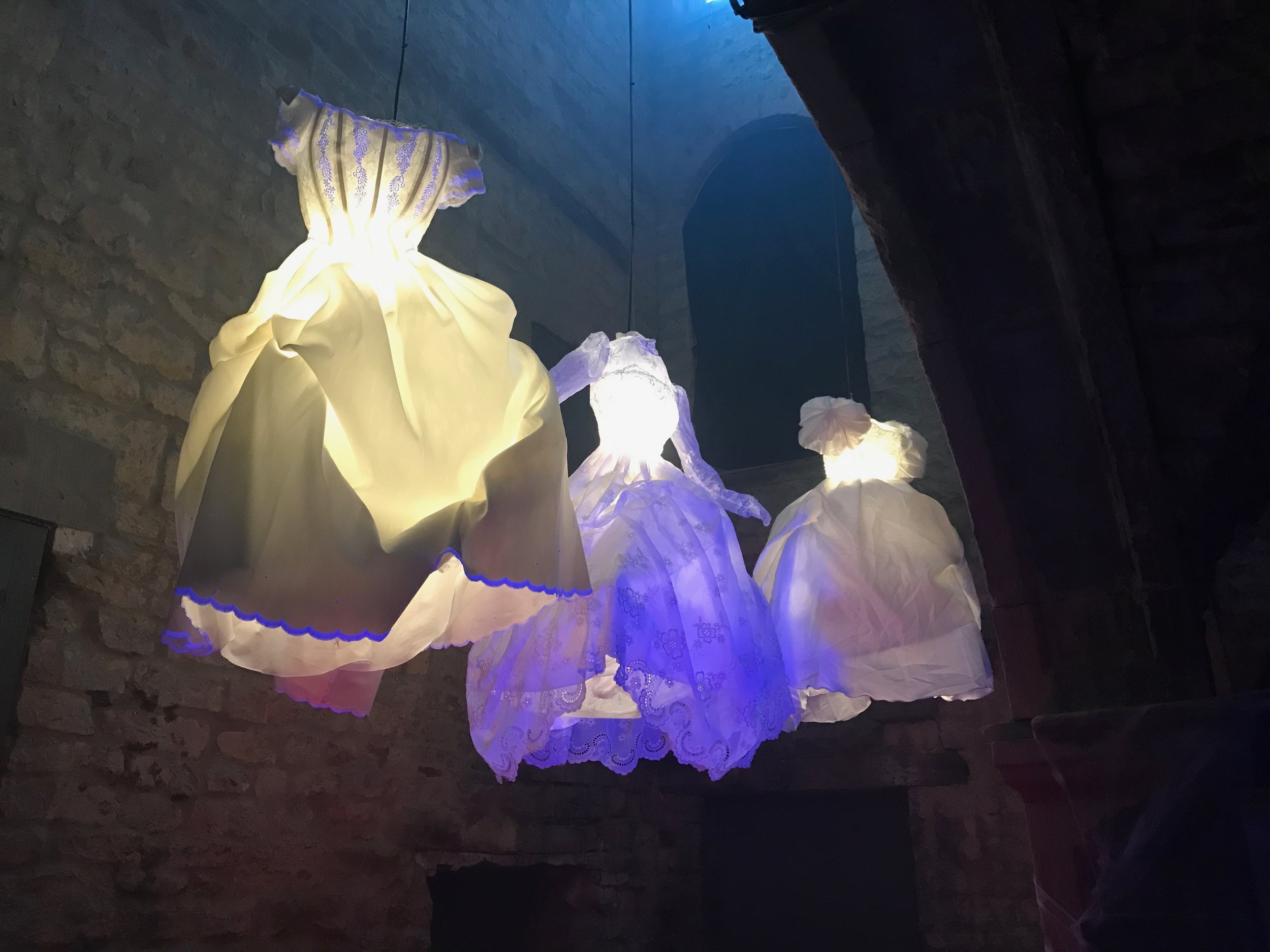 Illumination of silk or fabric dresses hanging on wires in a dimly lit space with brick walls, creating a glowing effect.