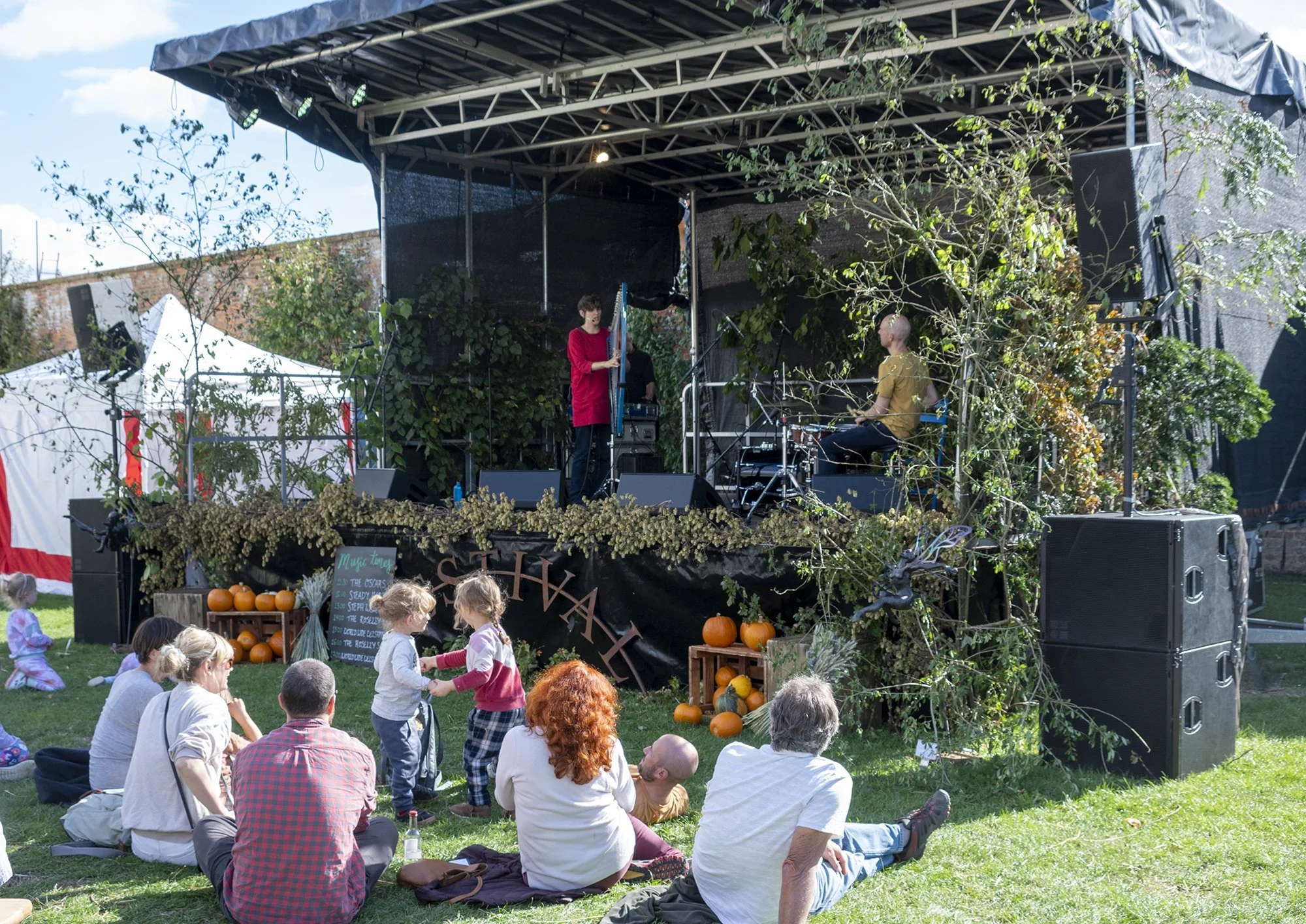 An outdoor stage decorated with pumpkins, gourds, and autumn foliage, hosting a musical performance during a fall festival. Children and adults are sitting on the grass, watching the performers and enjoying the event.