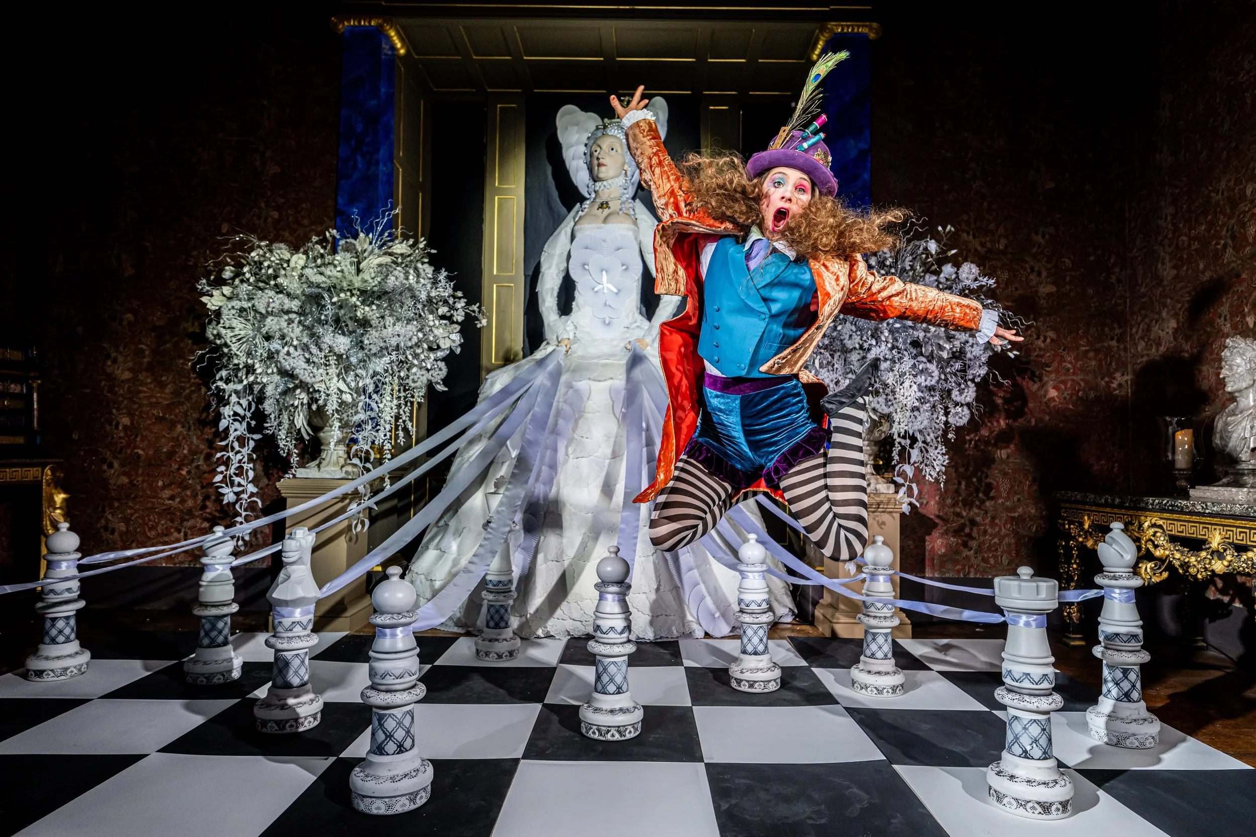 A theatrical display featuring a woman in colorful, eccentric costume with striped tights, a purple hat with peacock feathers, standing on a checkerboard floor, with a white dress and elaborate headpiece in the background.