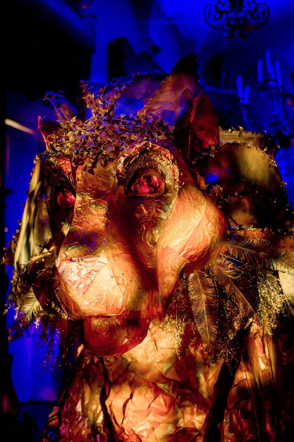 A colorful artistic sculpture of a human face made from crinkled paper, metallic leaves, and decorative elements, illuminated in dim blue and warm lighting.