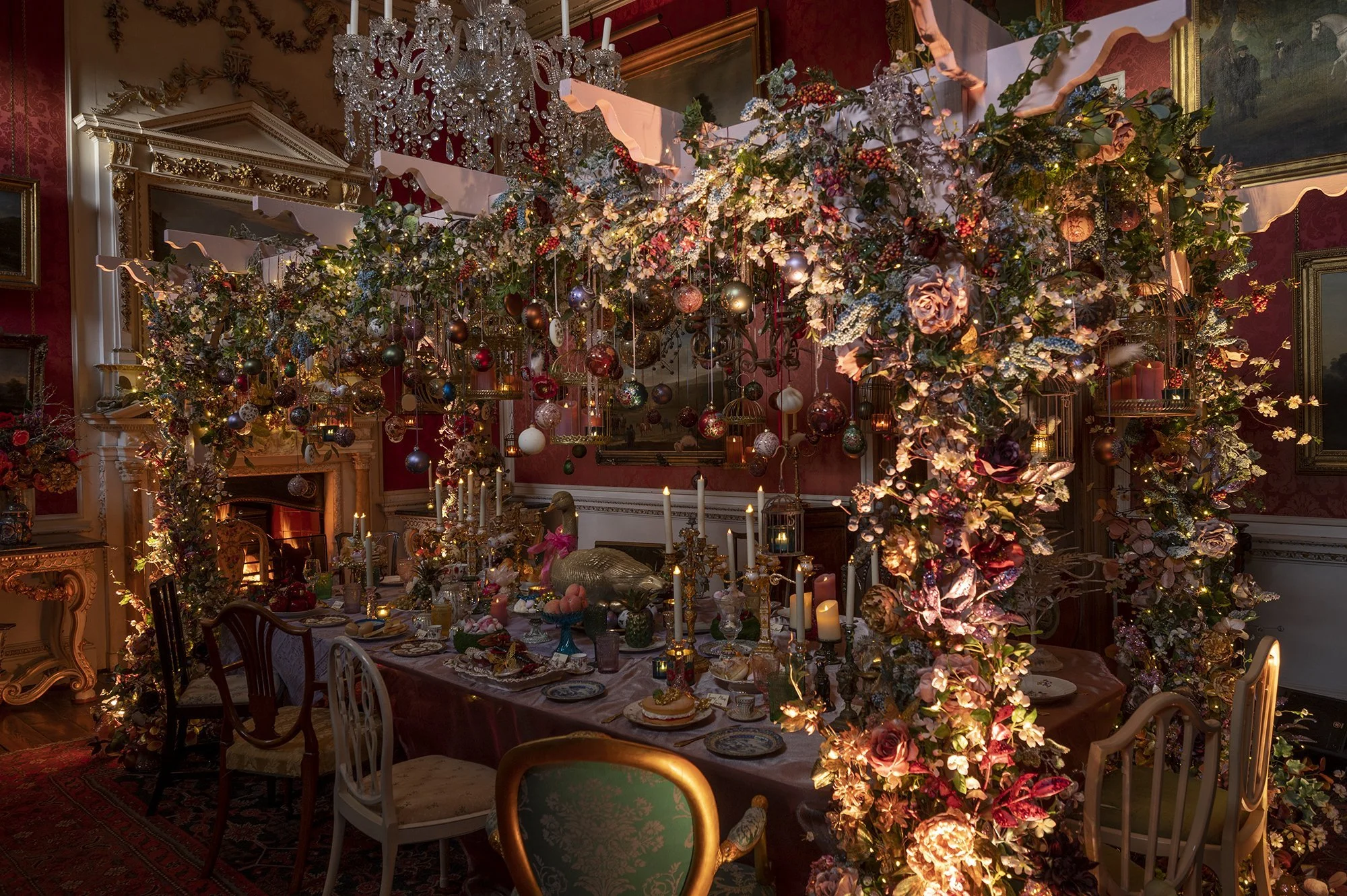 An ornate dining room decorated with a large floral and ornament chandelier, with a lavish table setting beneath it. The room has red walls, gilded picture frames, big windows with curtains, and a large painting on the wall. The table is set with var