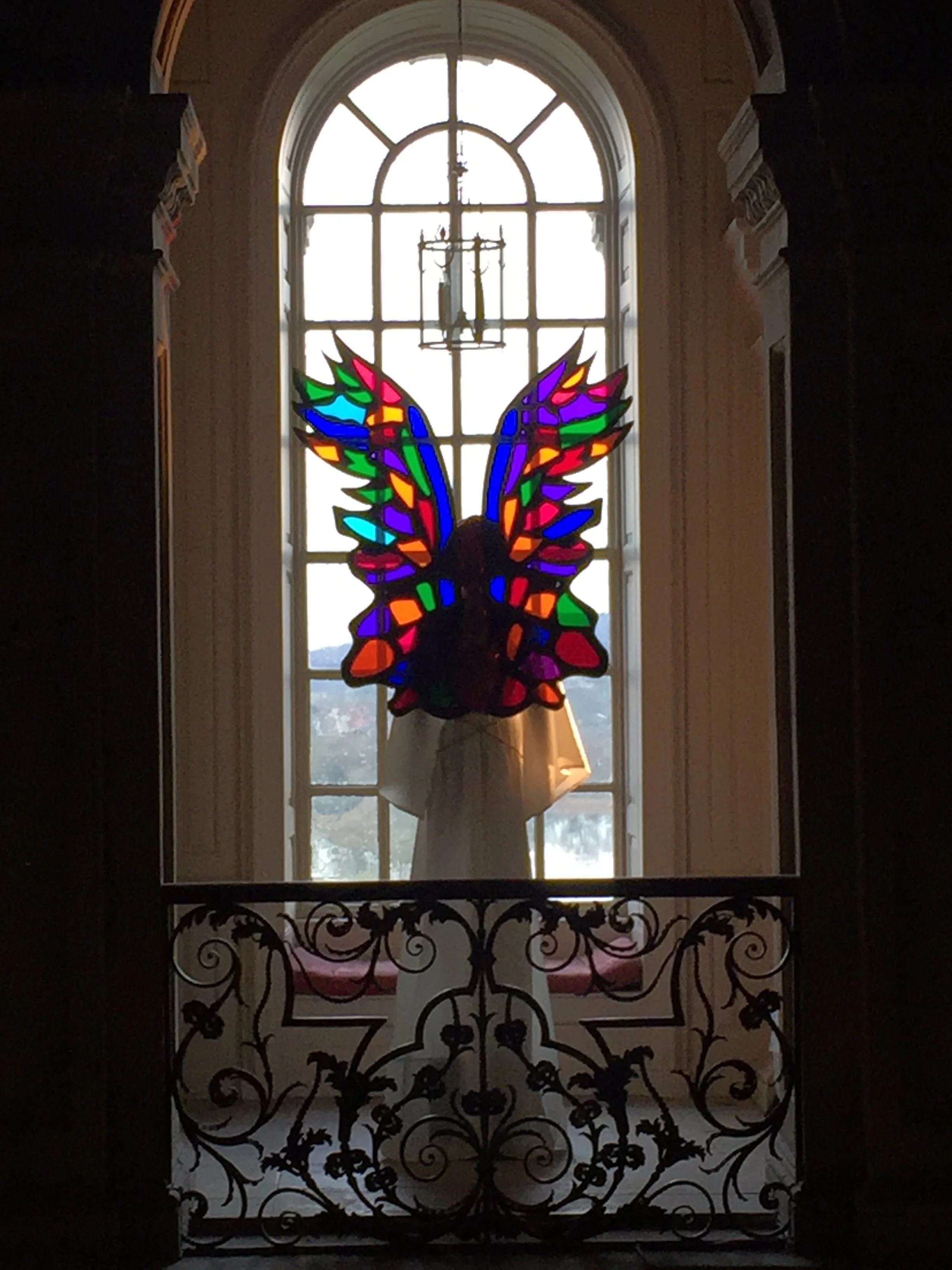 Colorful stained glass of a butterfly with wings spread, backlit by sunlight, with decorative wrought iron gate in front and a large arched window in the background.
