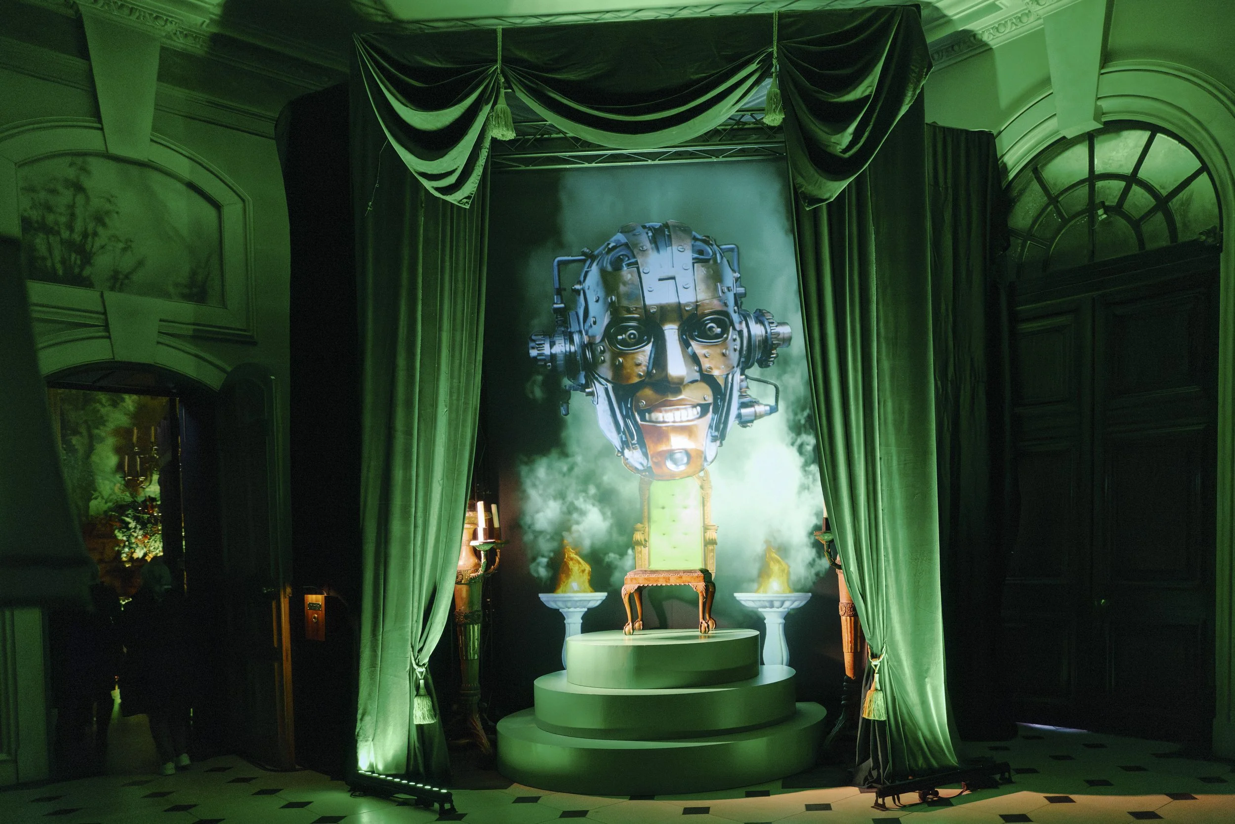 Stage with green curtains and a large robot head sculpture with metallic features and a big smile, surrounded by green lighting and fog, in a theatrical or exhibition setting.