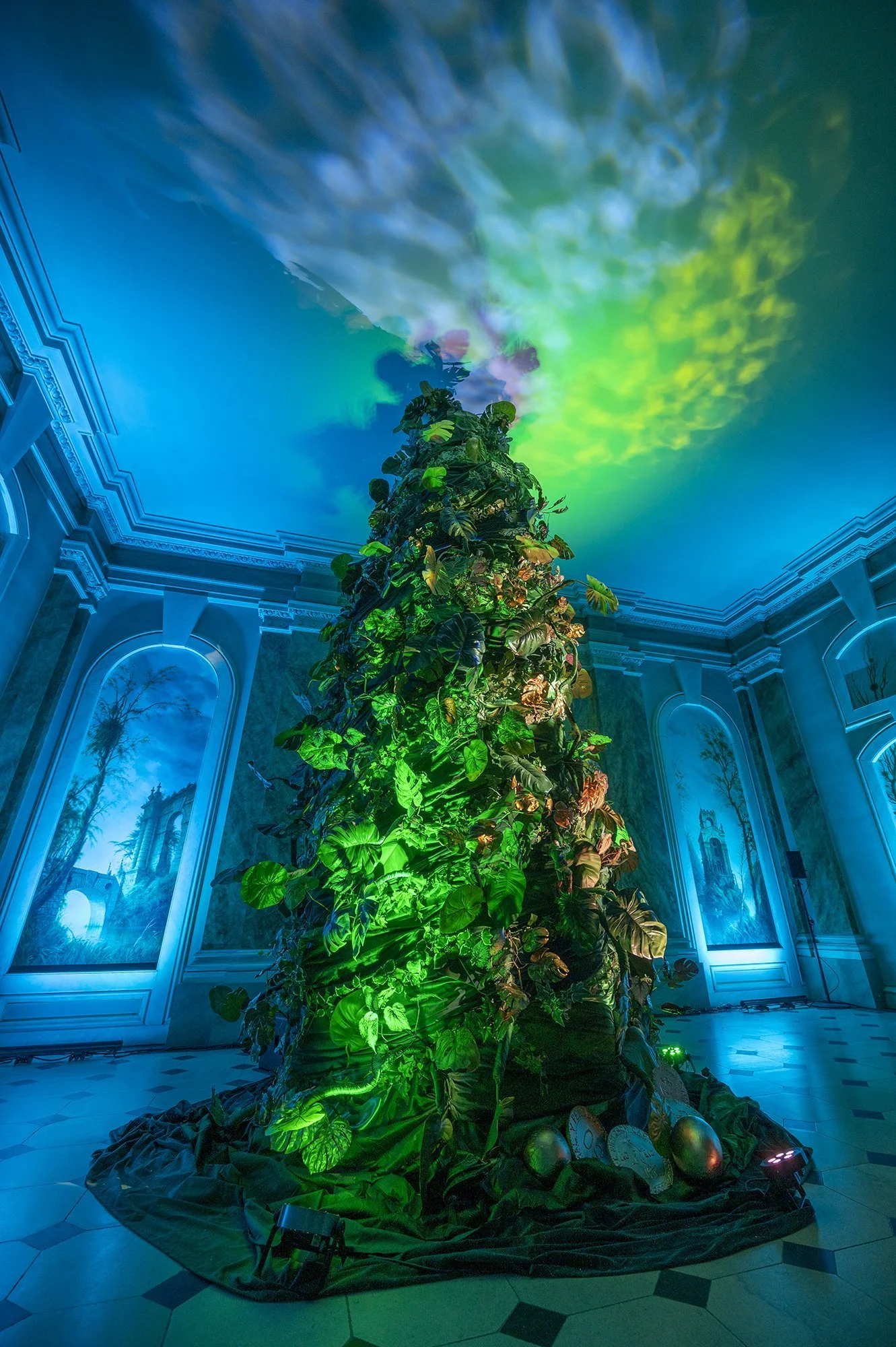 A large indoor plant display with leaves illuminated by green and purple lights, set against a decorated ceiling and walls with ornate moldings. The floor is tiled and there are stage lights on the floor.