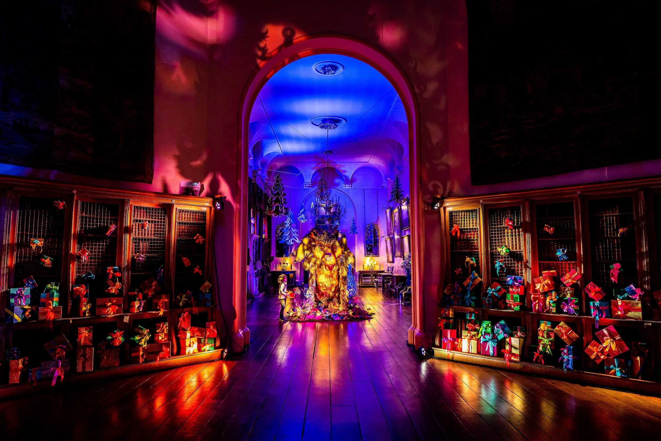 Decorated room with Christmas theme featuring colorful presents, illuminated holiday decorations, and a glowing sculpture central to the scene, with a blue lit ceiling and ornate architectural details.