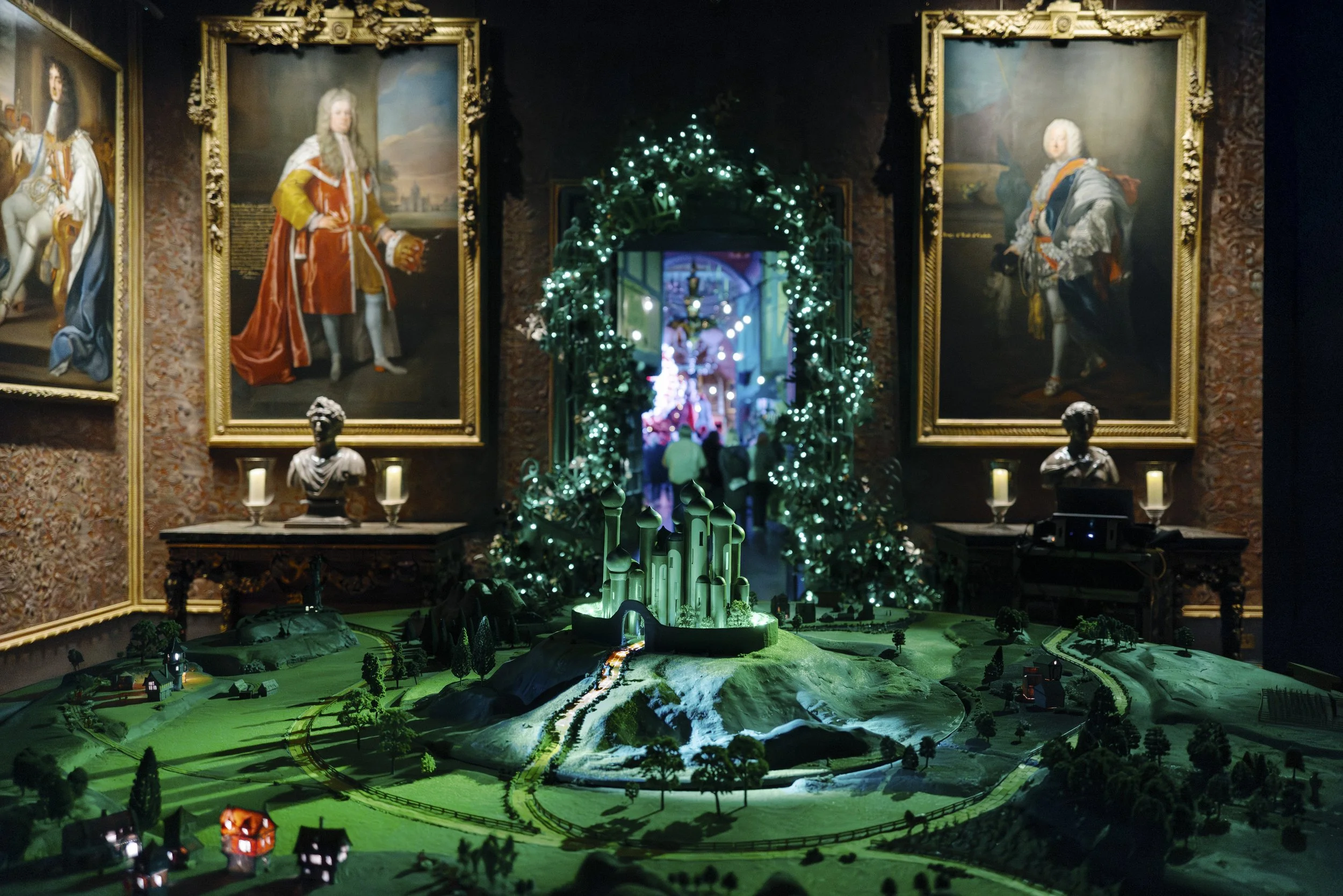 A diorama of a historical landscape with miniature buildings, roads, and trees illuminated by green and white lights, displayed indoors with framed portraits on the wall behind.