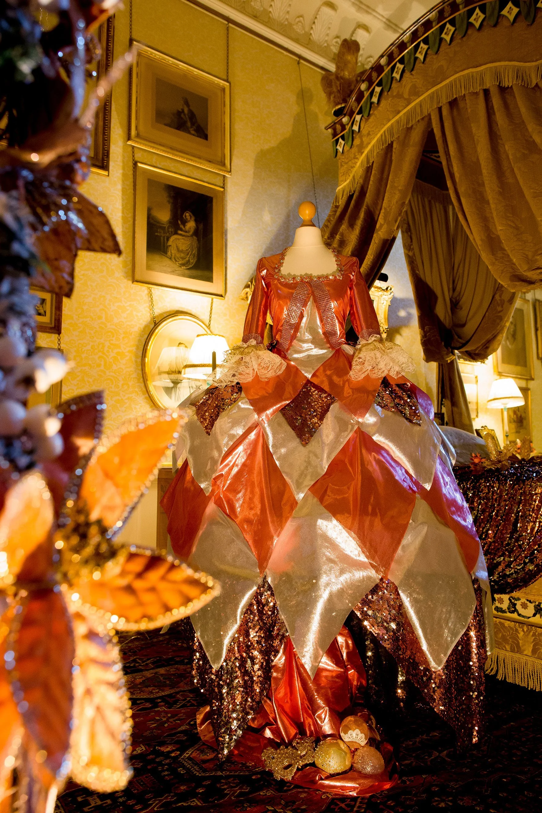 A doll dressed in an elaborate, colorful gown with gold and orange sequins, lace, and ruffles, displayed in a luxurious room with framed pictures on the wall, ornate curtains, and evening lighting.