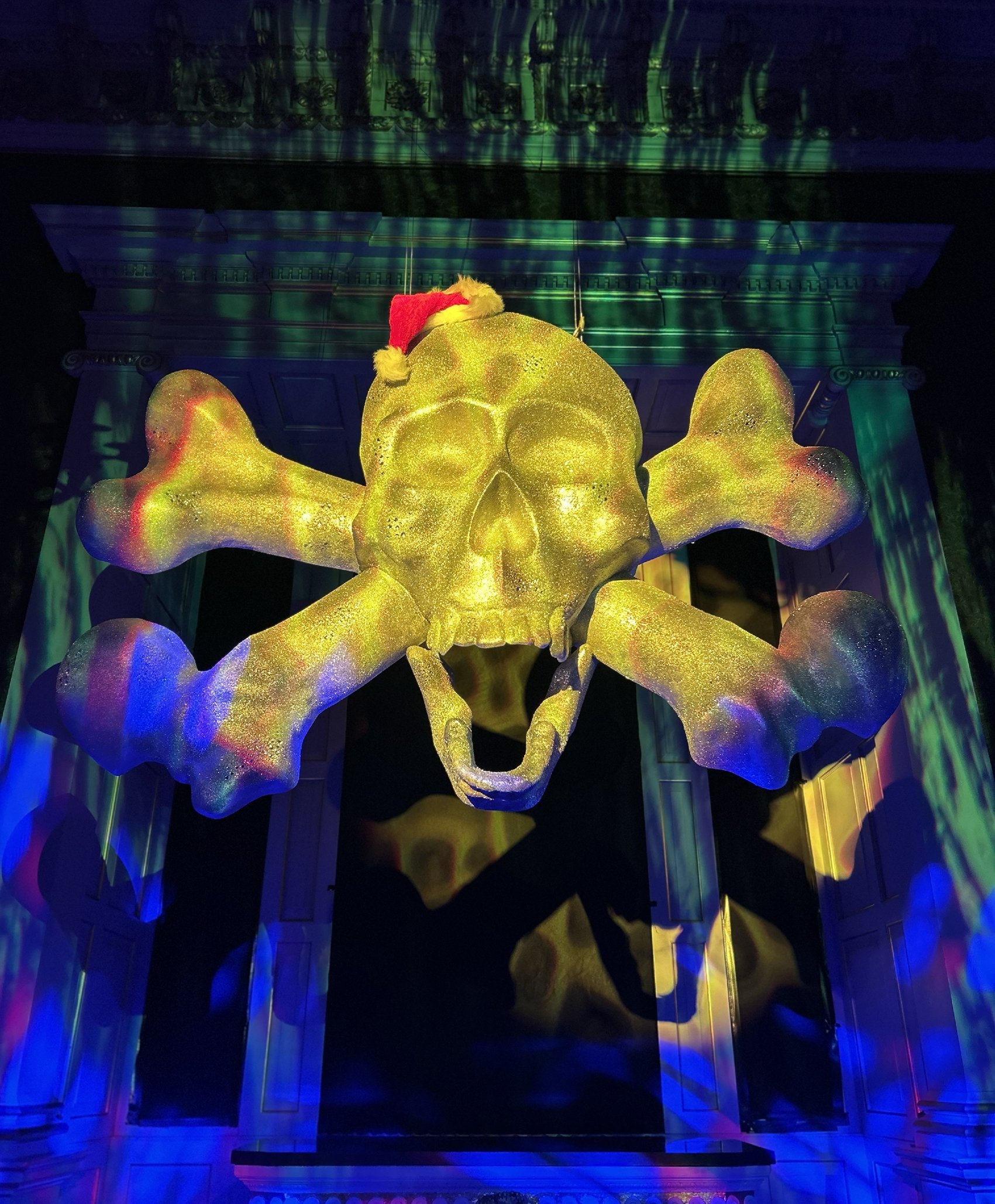 A large illuminated skull and butterfly sculpture with a Santa hat, set against a building with colorful lights reflected on its glass surface.