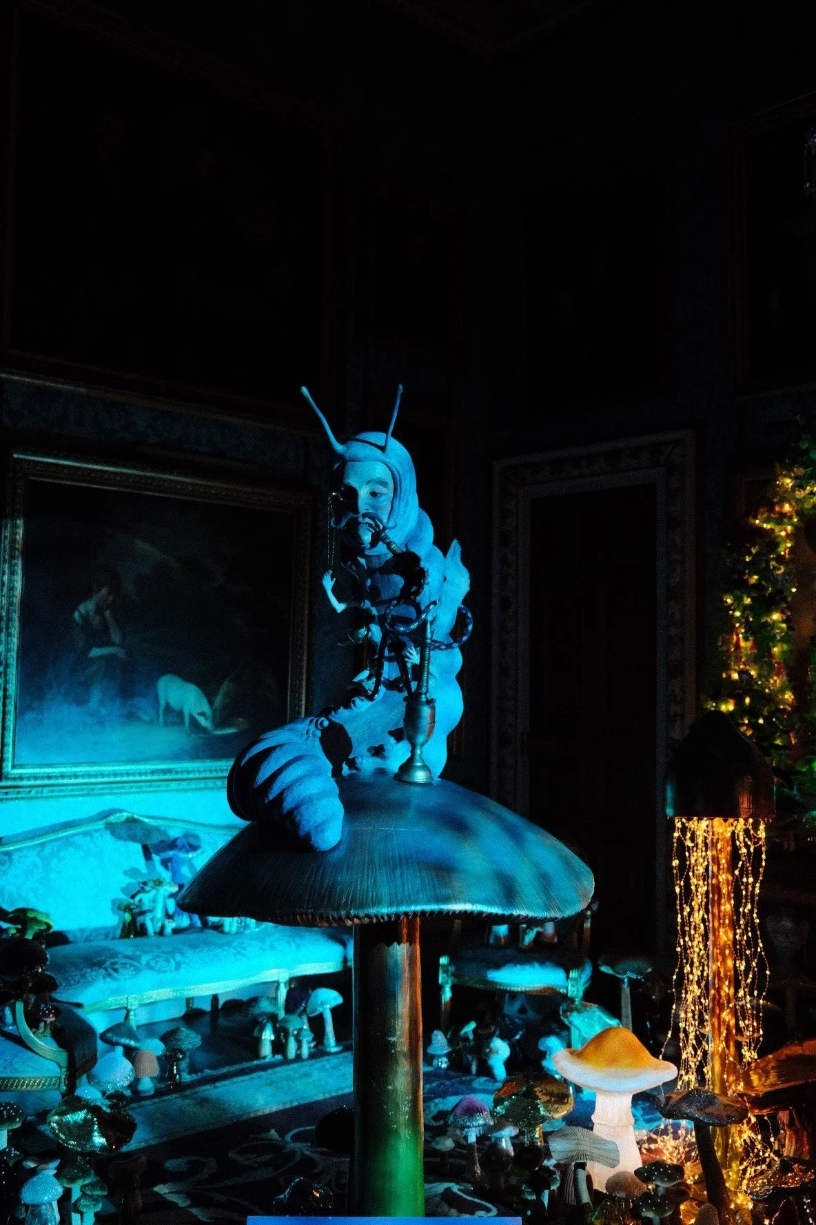 An artistic display featuring a sculpture of a human head with an insect-like face and antennas, fused with mechanical and organic elements, illuminated with blue lighting in a dimly lit room decorated with paintings, a Christmas tree, and various mu