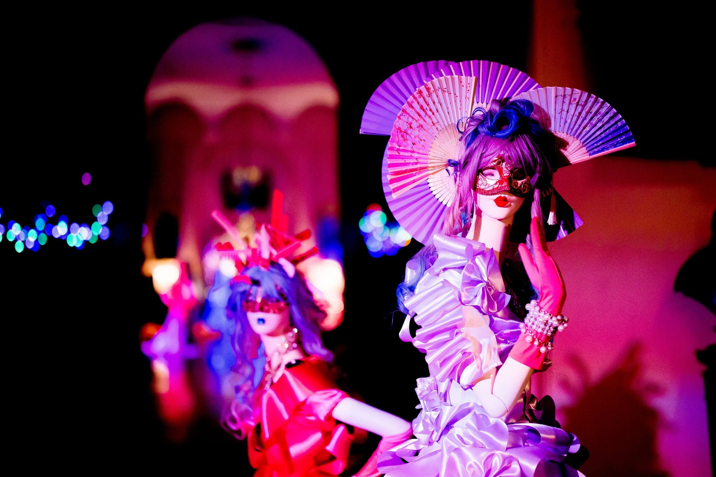 Colorful figurines with elaborate costumes, masks, and parasols, likely dolls or mannequins in a display, illuminated by vibrant pink, purple, and red lighting.