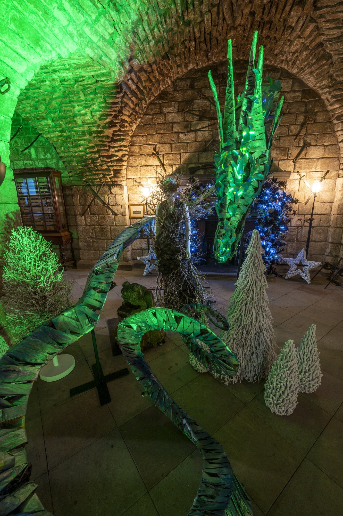 Indoor scene with decorative plants and illuminated sculptures, including a large green plant with glowing leaves, a white textured tree, and star-shaped lights, set against a brick arch ceiling background.