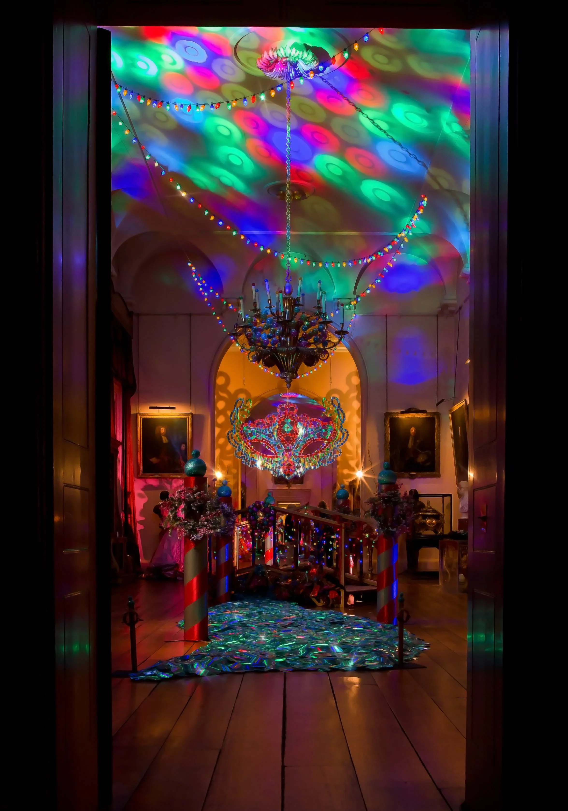 Colorful holiday decorations in a grand hallway including a chandelier, string lights, and confetti on the floor, with portraits on the walls.