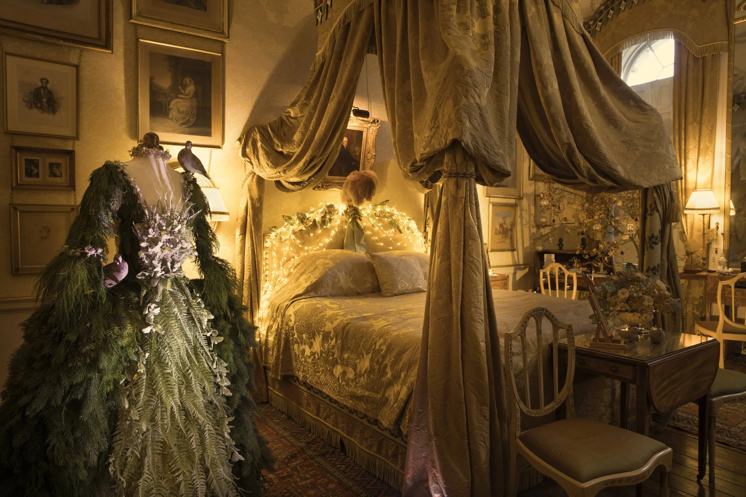 Vintage bedroom with ornate gold curtains, framed artwork on the walls, a decorated bed with fairy lights, a Christmas tree with ornaments, and antique furniture.
