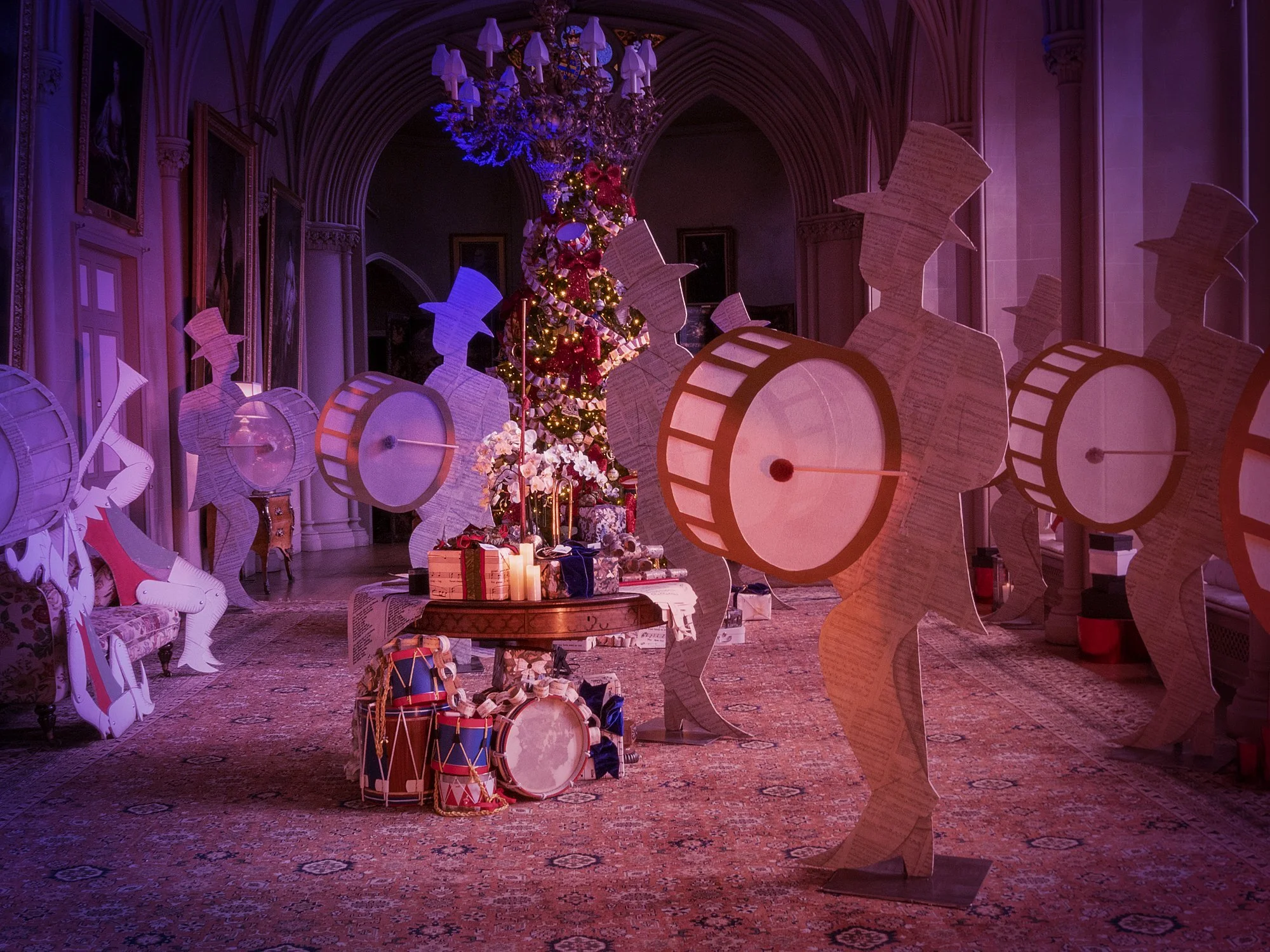 A decorated Christmas tree with red, gold, and white ornaments and lights, surrounded by cardboard cutouts of band members playing musical instruments, inside a grand hall with high arched ceilings and ornate details.