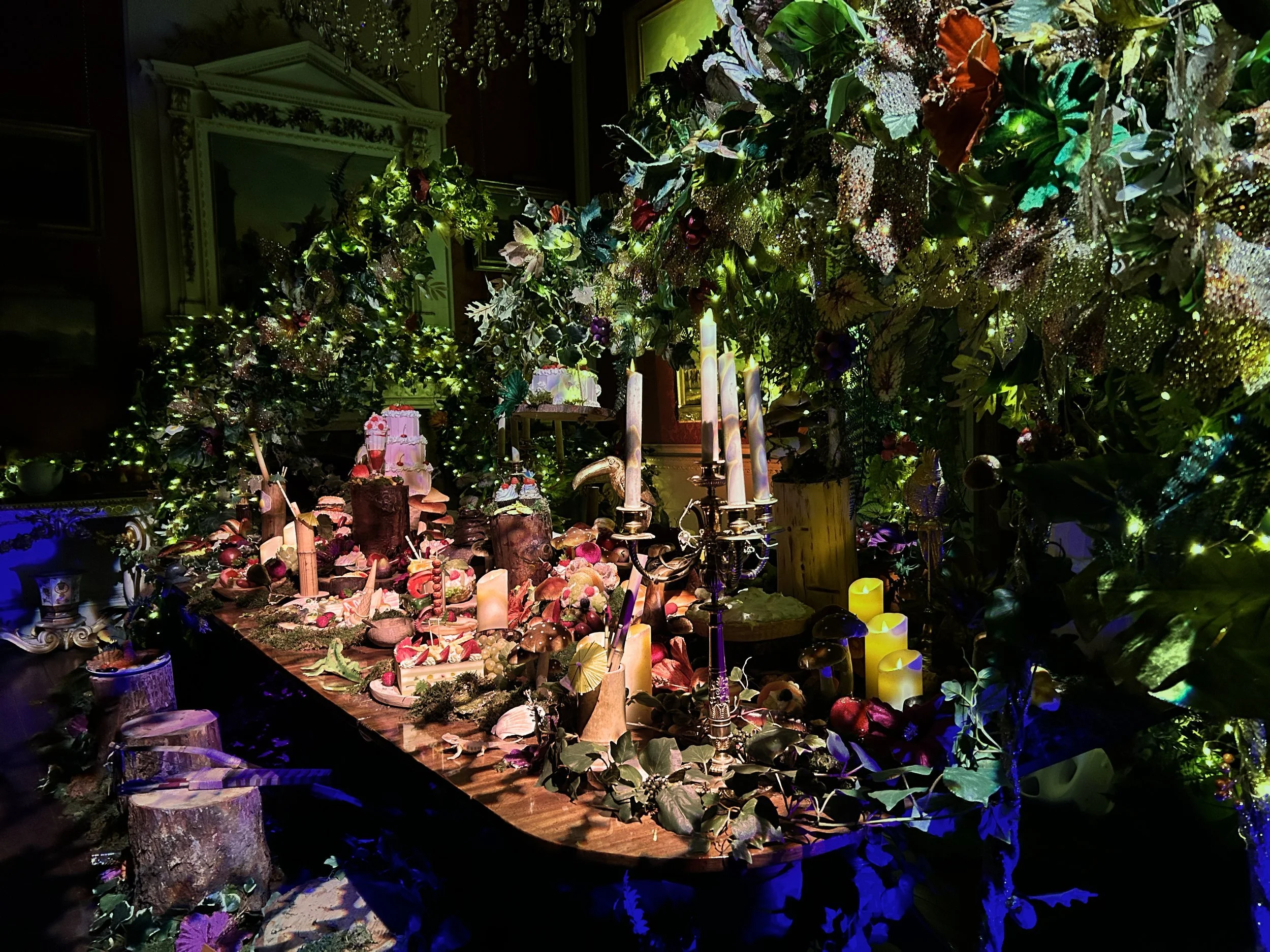 A decorated table with candles, desserts, and greenery, surrounded by a lush green and gold floral arch and dim ambient lighting.