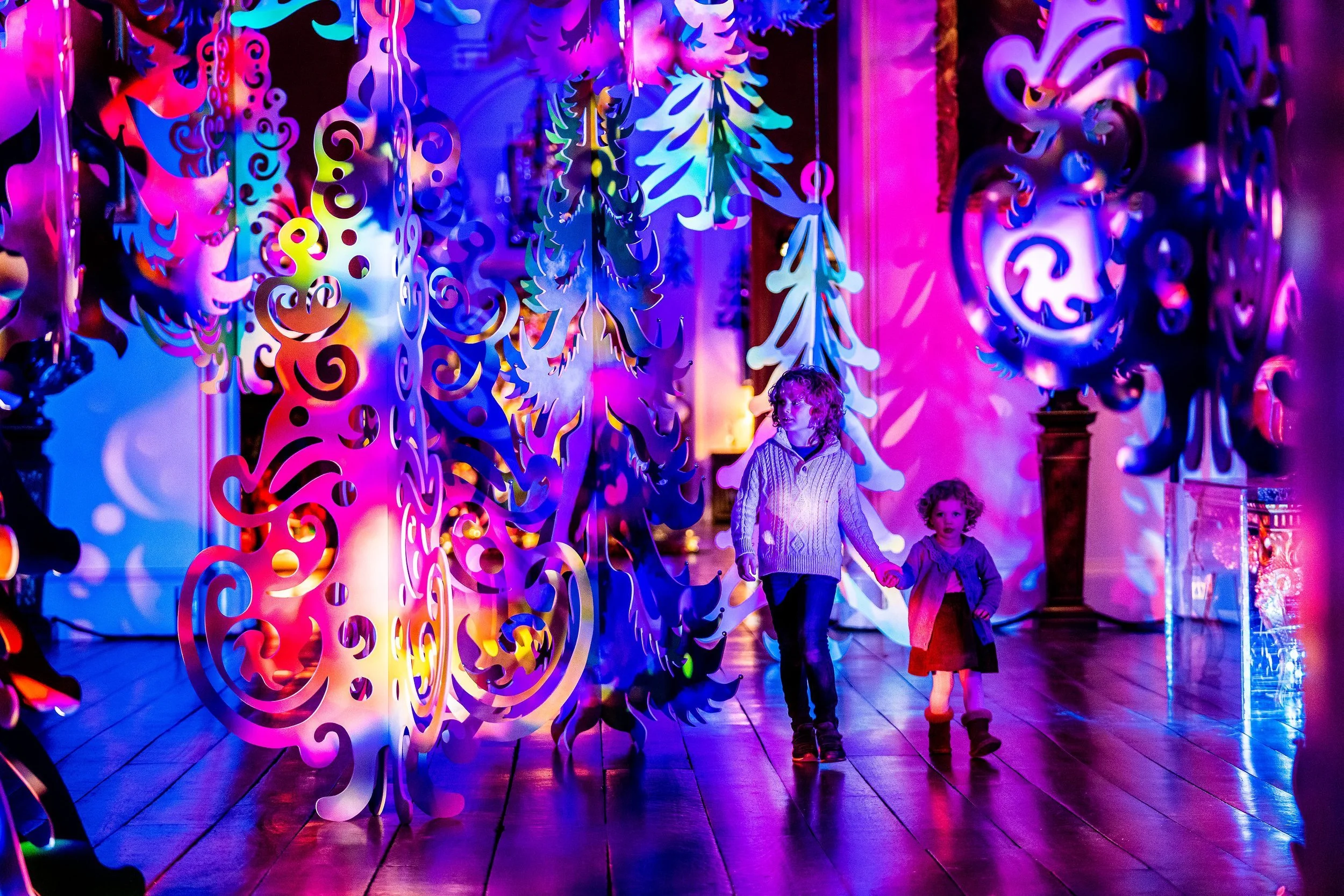 Children walking through an art installation of large, colorful, swirling shapes illuminated with vibrant pink, purple, blue, and yellow lights in a gallery or museum setting.