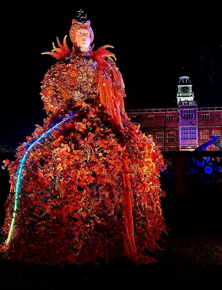 A large, colorful floral sculpture of a woman wearing a crown, illuminated at night with a backdrop of a brick building and a clock tower.
