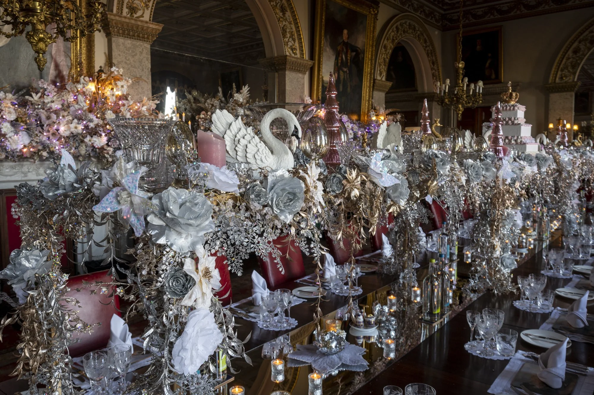 A luxurious banquet table with ornate decorations including silver and pink glassware, white swan figurines, candles, and floral arrangements, all set in a grand, gold-accented room with classical paintings.