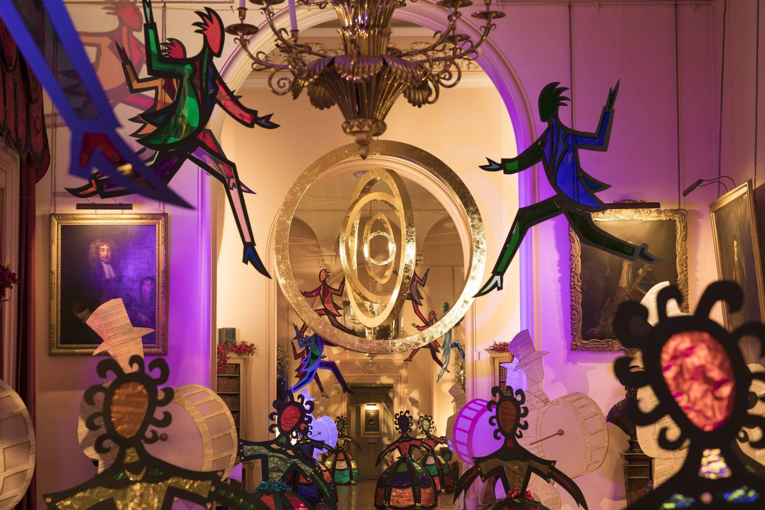Decorative art installation in an elegant room with ornate chandelier, paintings, and floral arrangements. The installation features colorful cutouts of figures and geometric shapes hanging from the ceiling and placed in the room.