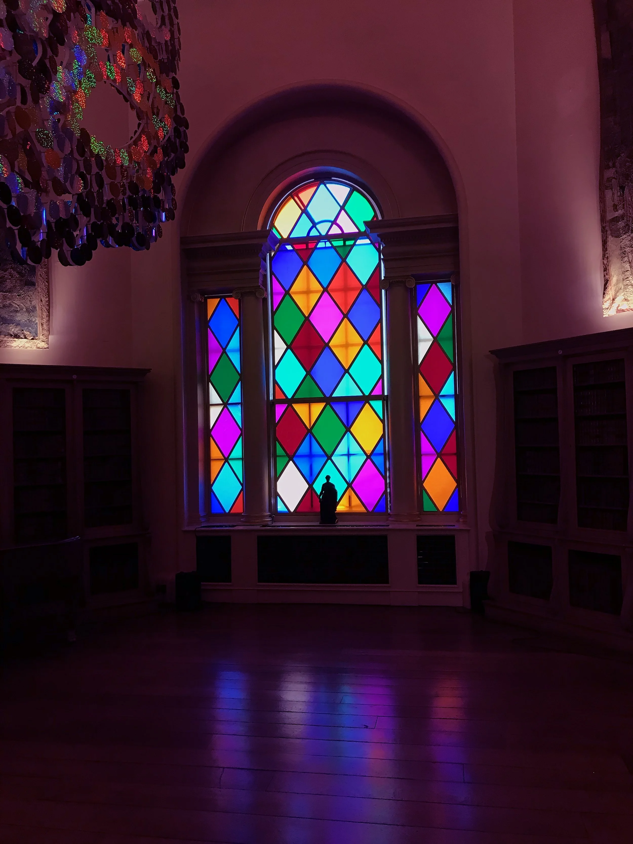 Colorful stained glass window with geometric patterns, illuminated in a dim room, a small silhouette figurine at the center.