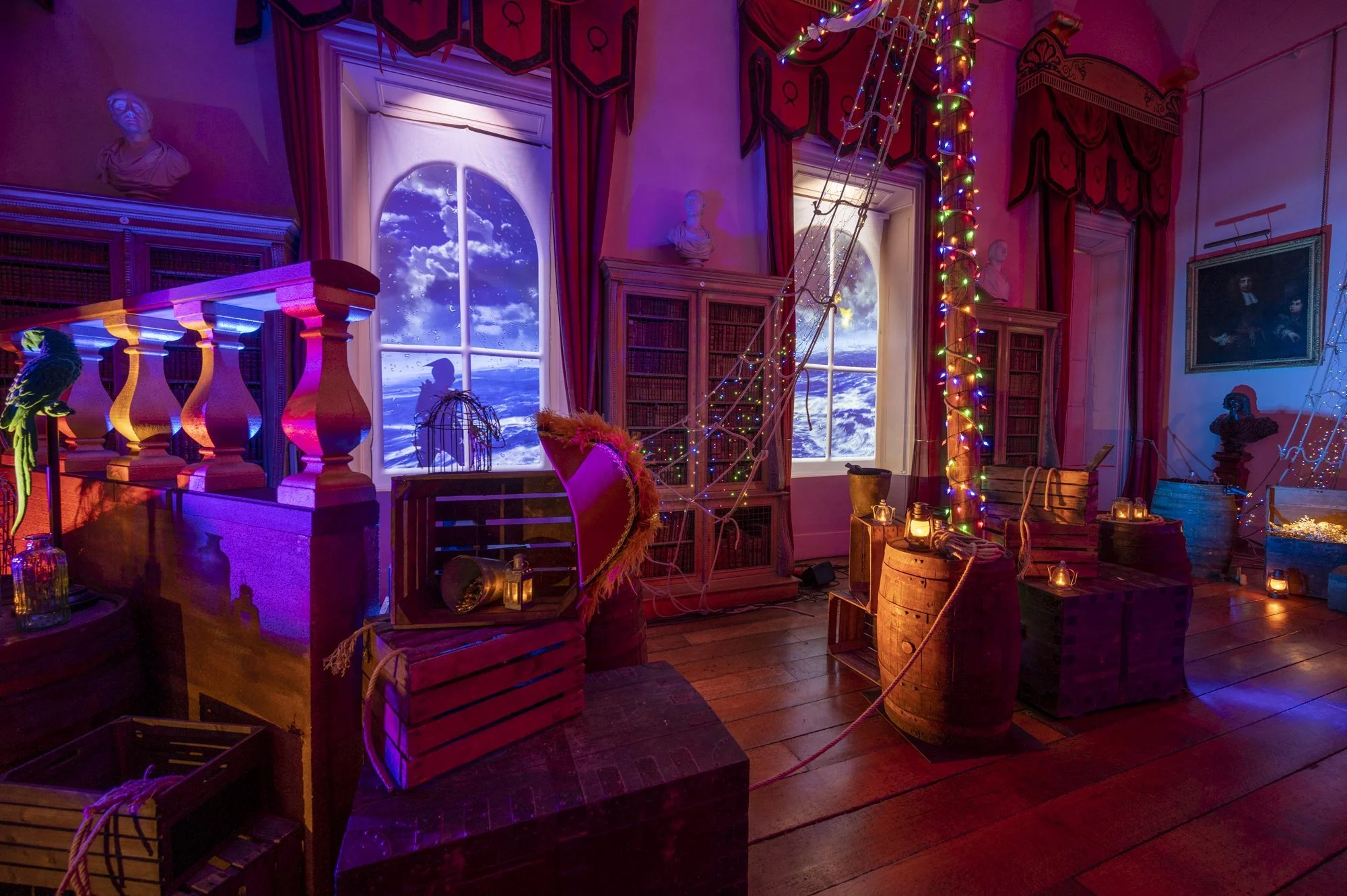 A room decorated with colorful string lights, wooden sculptures, bookshelves, framed pictures, and windows showing a moonlit sky with clouds. The room has a vintage and whimsical atmosphere with various objects like lanterns, a birdcage, and crates.