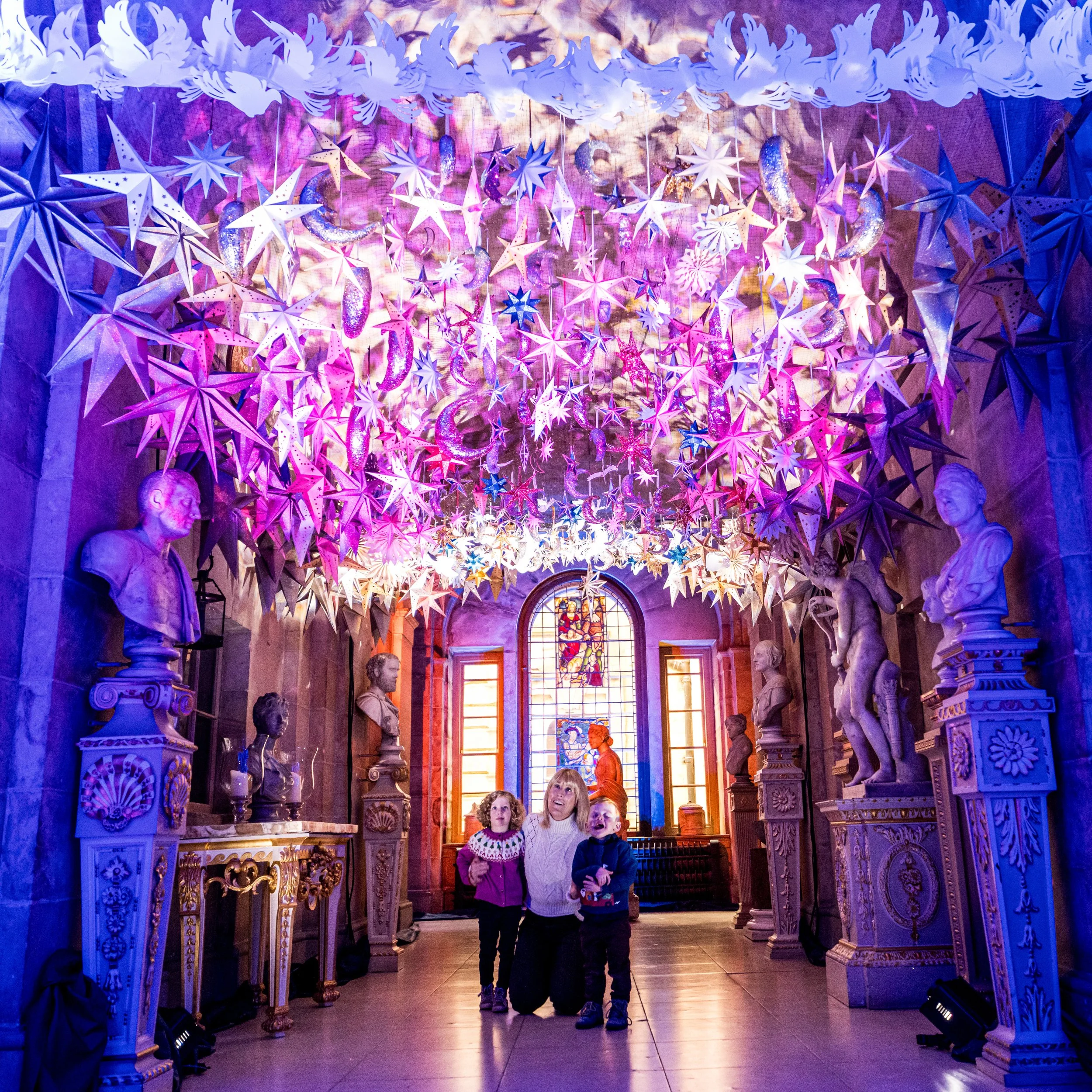 Three children inside a cathedral with ornate columns and statues, looking up at suspended star and moon decorations with colored lighting.