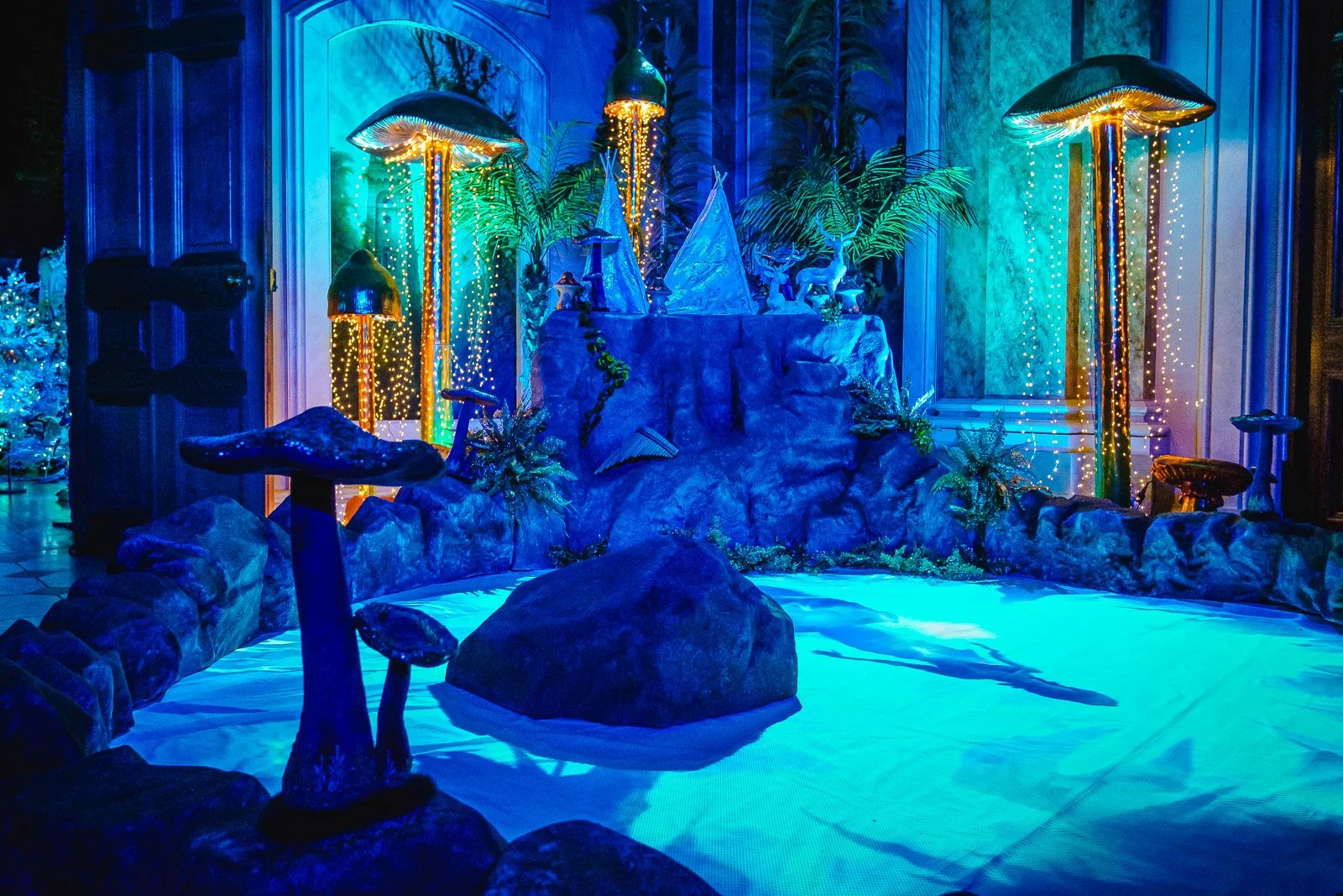 A fantasy-themed indoor landscape with large mushrooms, rocks, and lush plants illuminated by colorful lights, creating an enchanted forest scene.