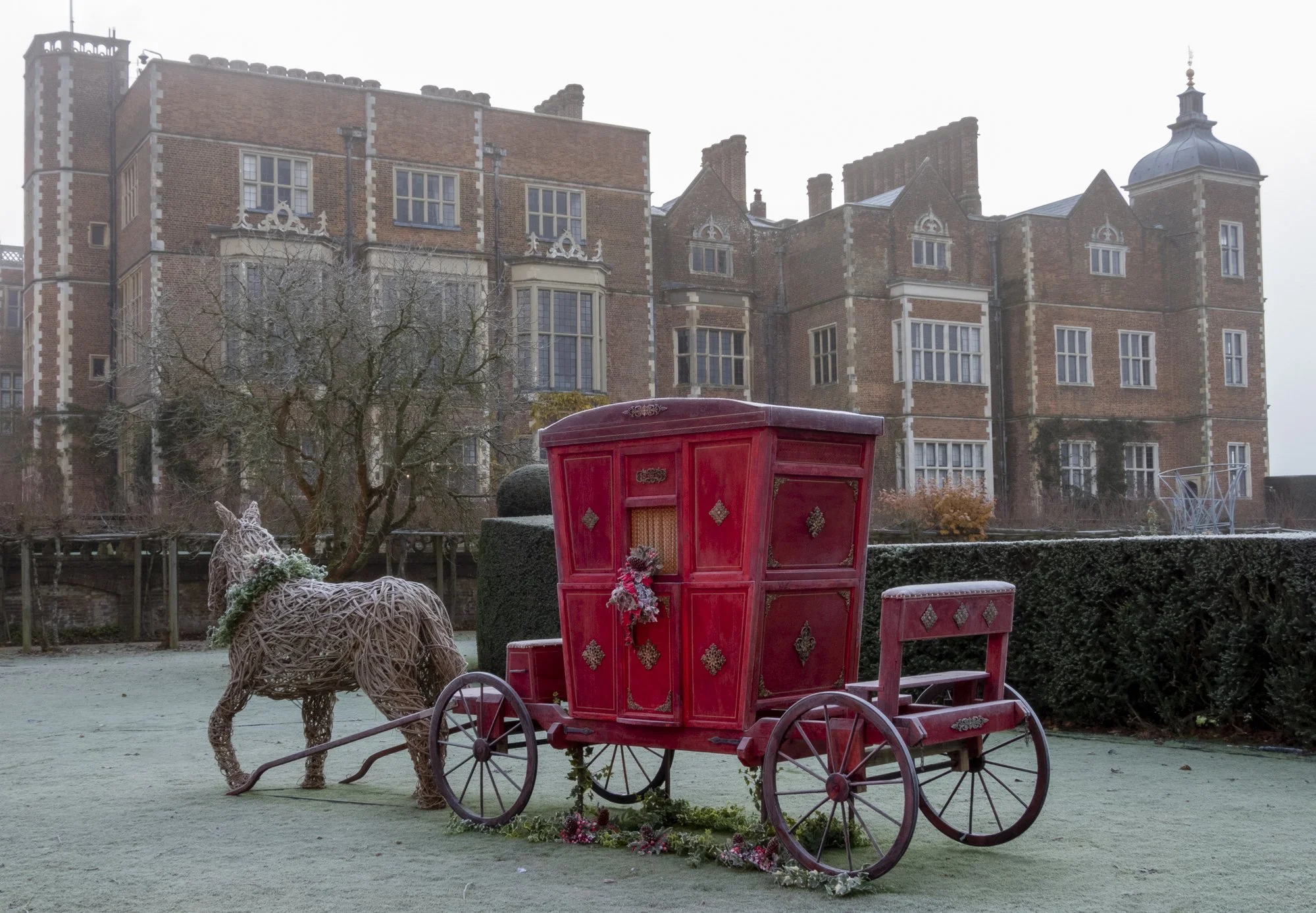 A red antique-style horse-drawn carriage with intricate gold detailing, decorated with holiday ornaments, is parked outdoors on a frosty ground. In the background, there is a leafless tree, a life-sized wire reindeer, and a historic brick building wi