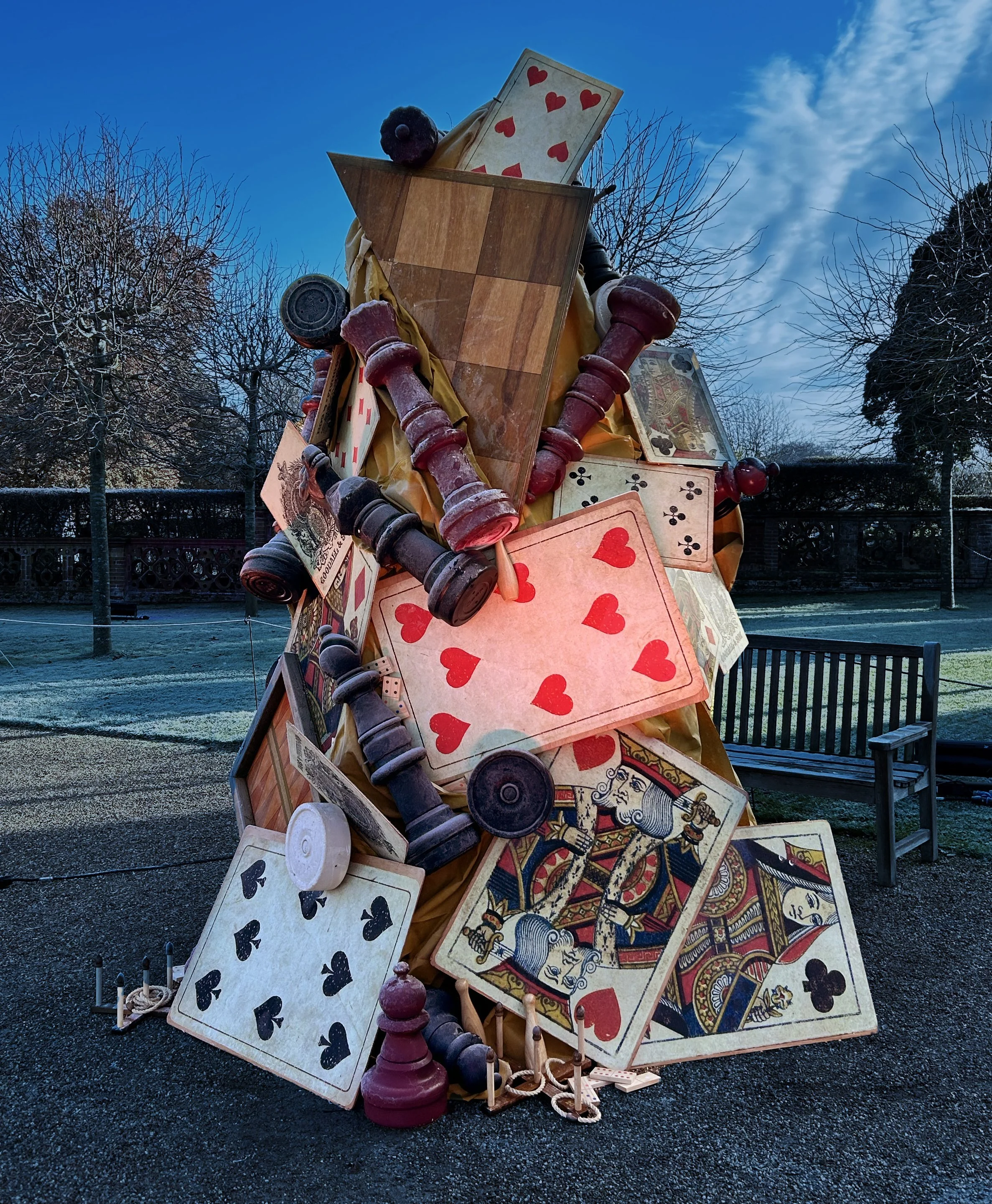 Sculpture made of oversized playing cards, chess pieces, and game pieces outdoors during daytime.