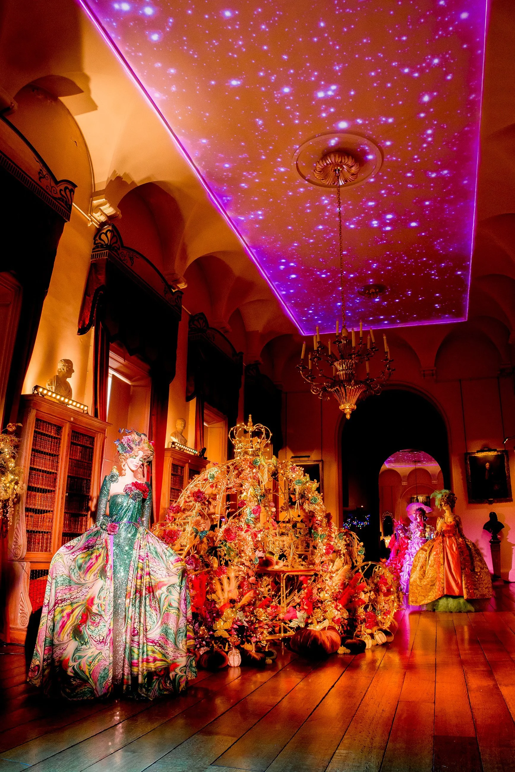 Colorful mannequins dressed in elaborate, vibrant, floral-patterned gowns on display in an ornate room with a starry ceiling projection, chandeliers, and wooden floors.