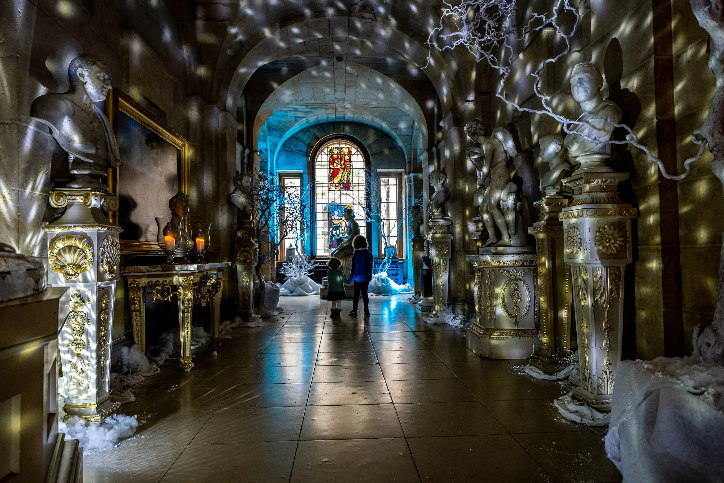 A grand hallway with marble bust sculptures lining the walls, a stained glass window depicting figures, and snow and ice scattered across the floor.