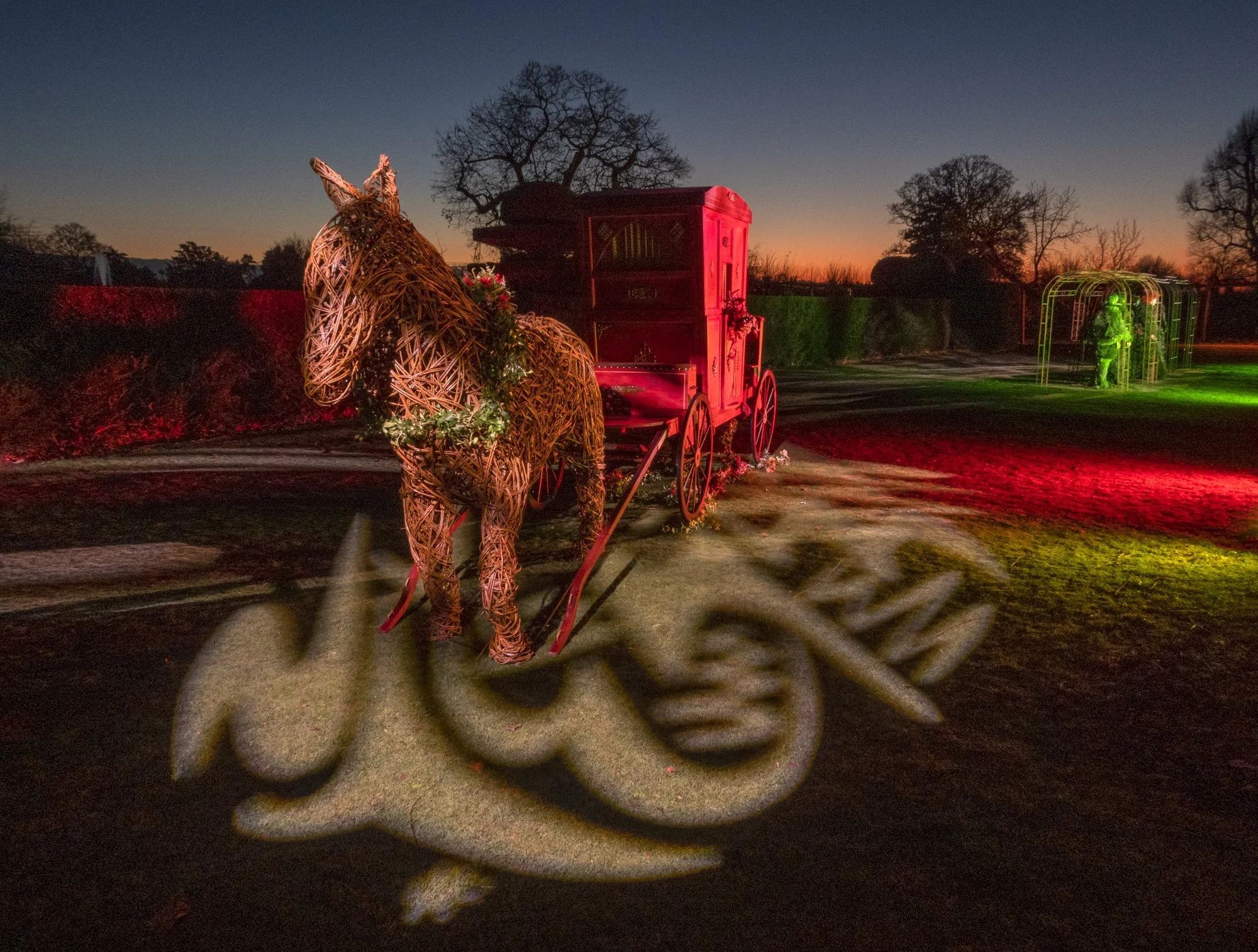 Decorative scene with a wicker rabbit and a red carriage, illuminated by colorful lights, set outdoors at dusk with trees in the background.
