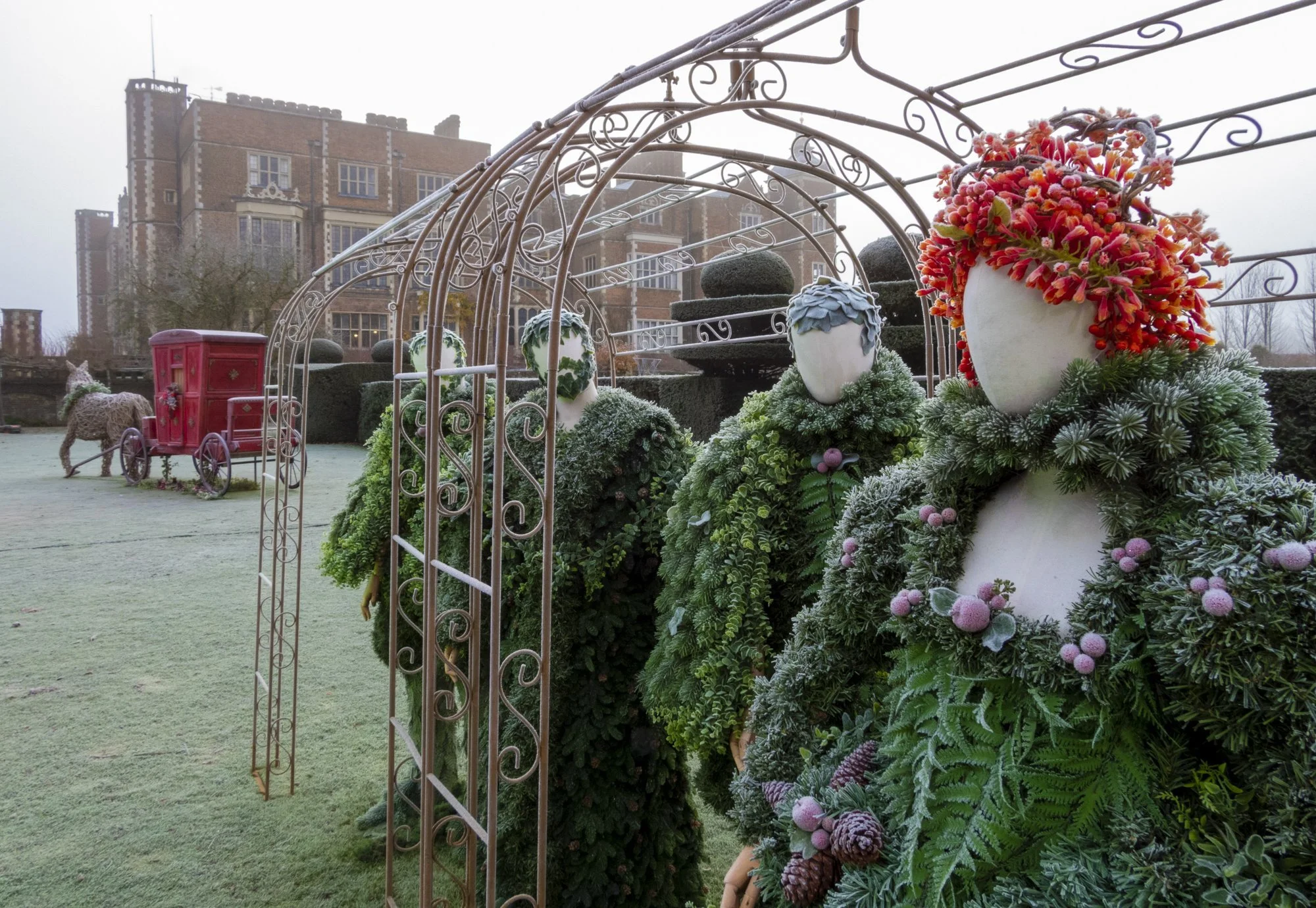 A garden scene with Christmas decorations, including a red sleigh, decorated with a bow, and a deer figure. There is an ornate metal archway decorated with large white vases filled with pink and red flowers and evergreen branches. In the background, 