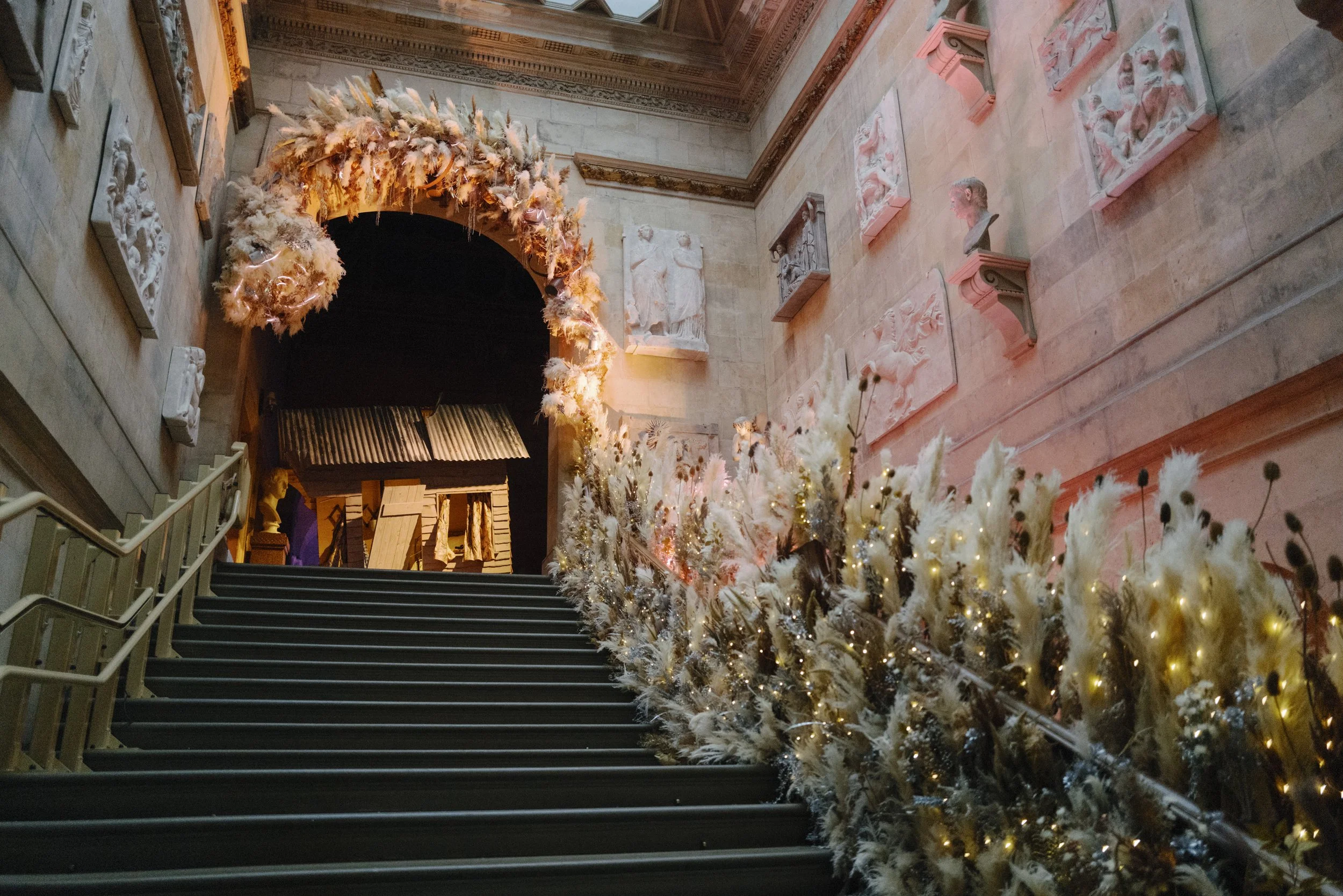 A decorated staircase with white Christmas trees and ornaments, leading up to an archway adorned with feathers and warm lighting inside a historic building with detailed wall sculptures.