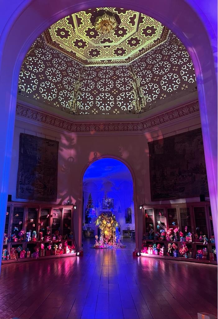 An elaborately decorated room with a lit dome ceiling projecting intricate patterns. The room features a central display or sculpture illuminated with vibrant colors, surrounded by shelves filled with wrapped gifts or decorations. The lighting create